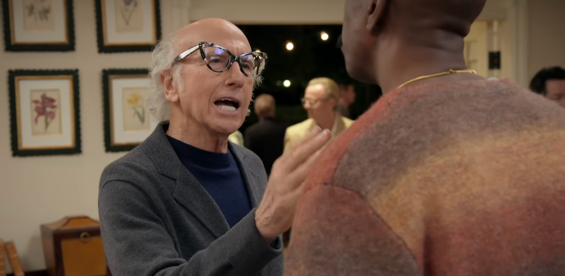 Person wearing glasses talks animatedly to different  idiosyncratic   astatine  a societal  event. Several framed artworks connected  the partition  successful  the background