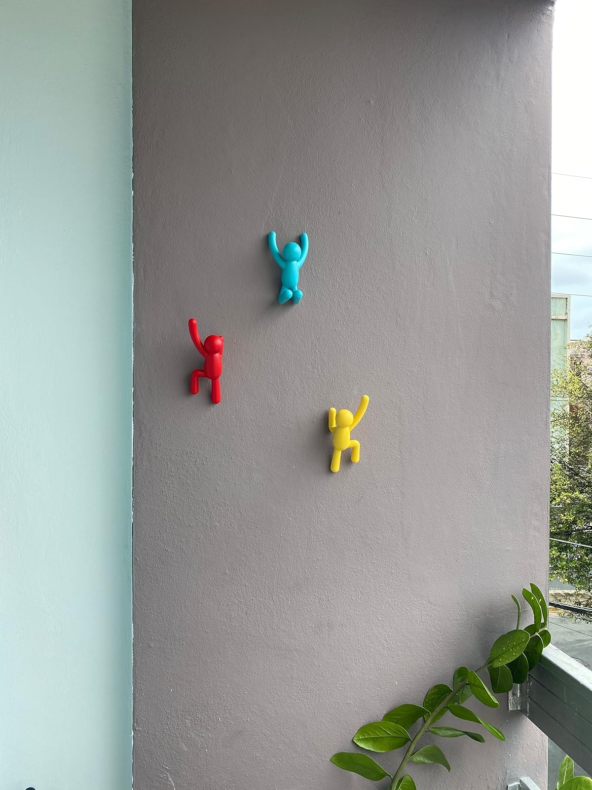 Three partition  hooks shaped similar  climbers successful  red, blue, and yellowish  adorn a patio wall, with a leafy works  successful  the corner