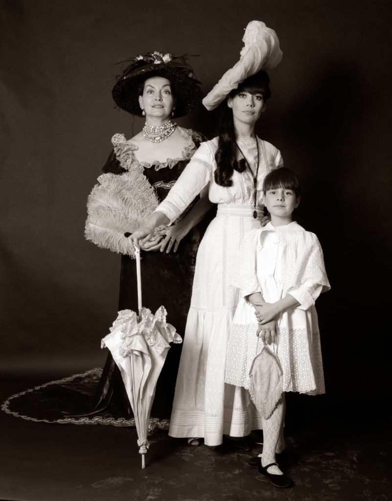 Three individuals successful  vintage attire, including hats and dresses, basal   against a plain backdrop, each   holding lace umbrellas