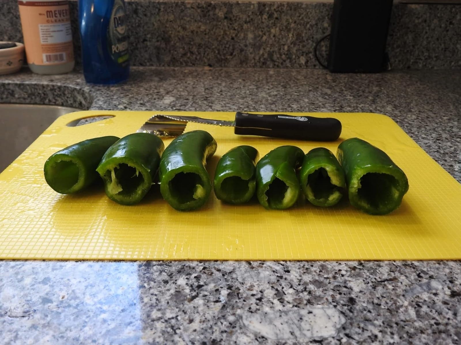 Six halved jalapeños with seeds removed, arranged connected  a yellowish  cutting committee  connected  a granite countertop. Peeler with a achromatic  grip  nearby