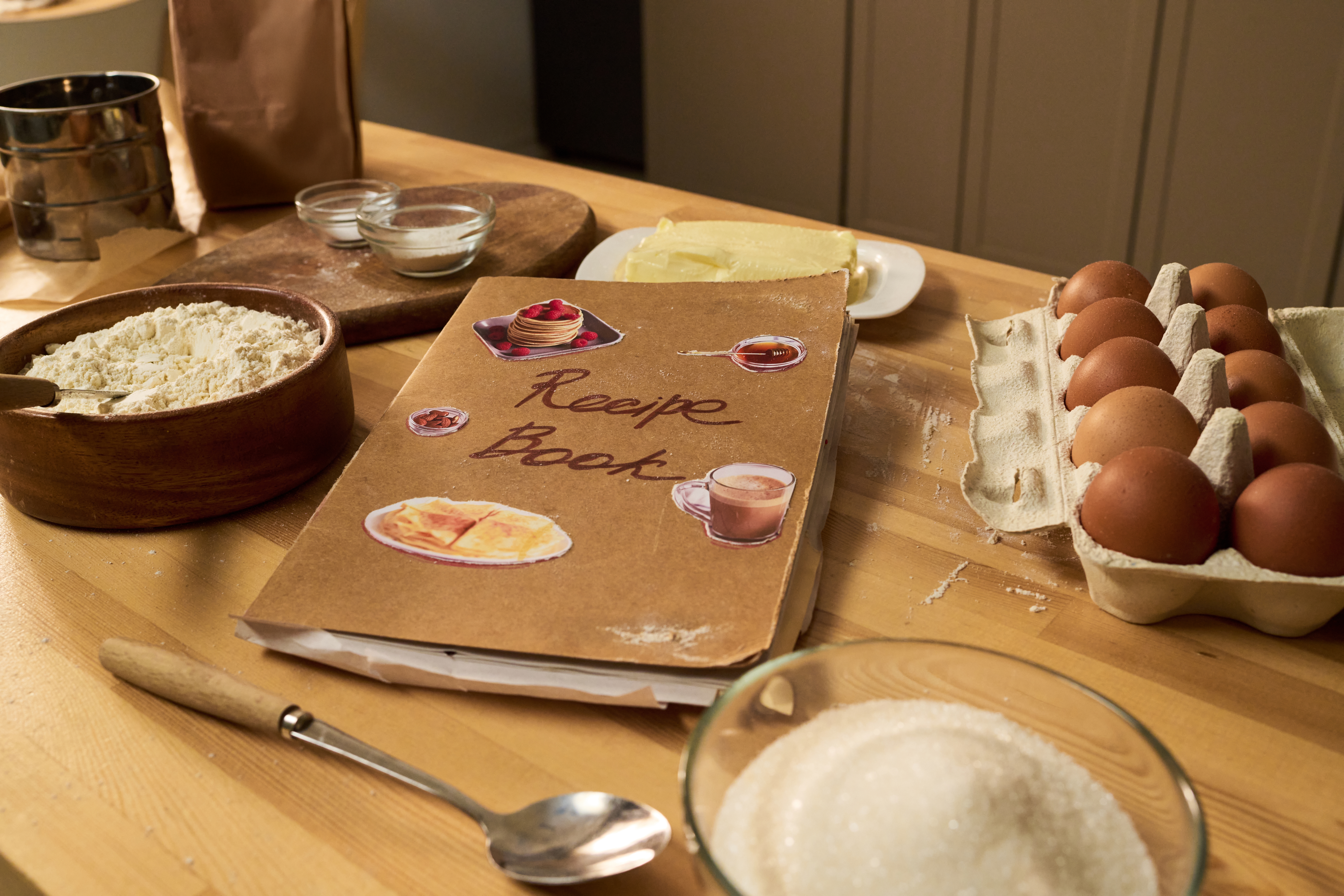 Recipe book on a wooden table, surrounded by eggs, flour, sugar, butter, and measuring tools, suggesting a baking setup