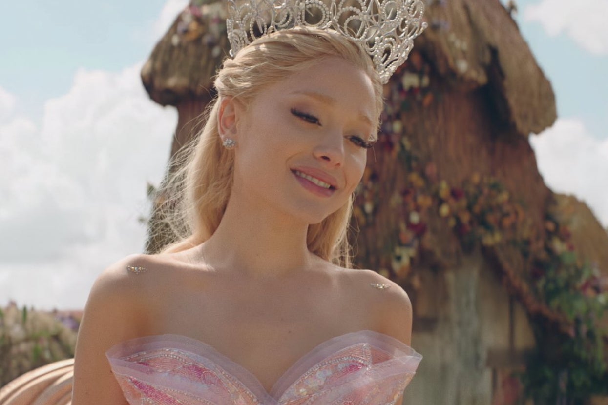 Person in a flowing gown and ornate crown, smiling outdoors by a rustic structure