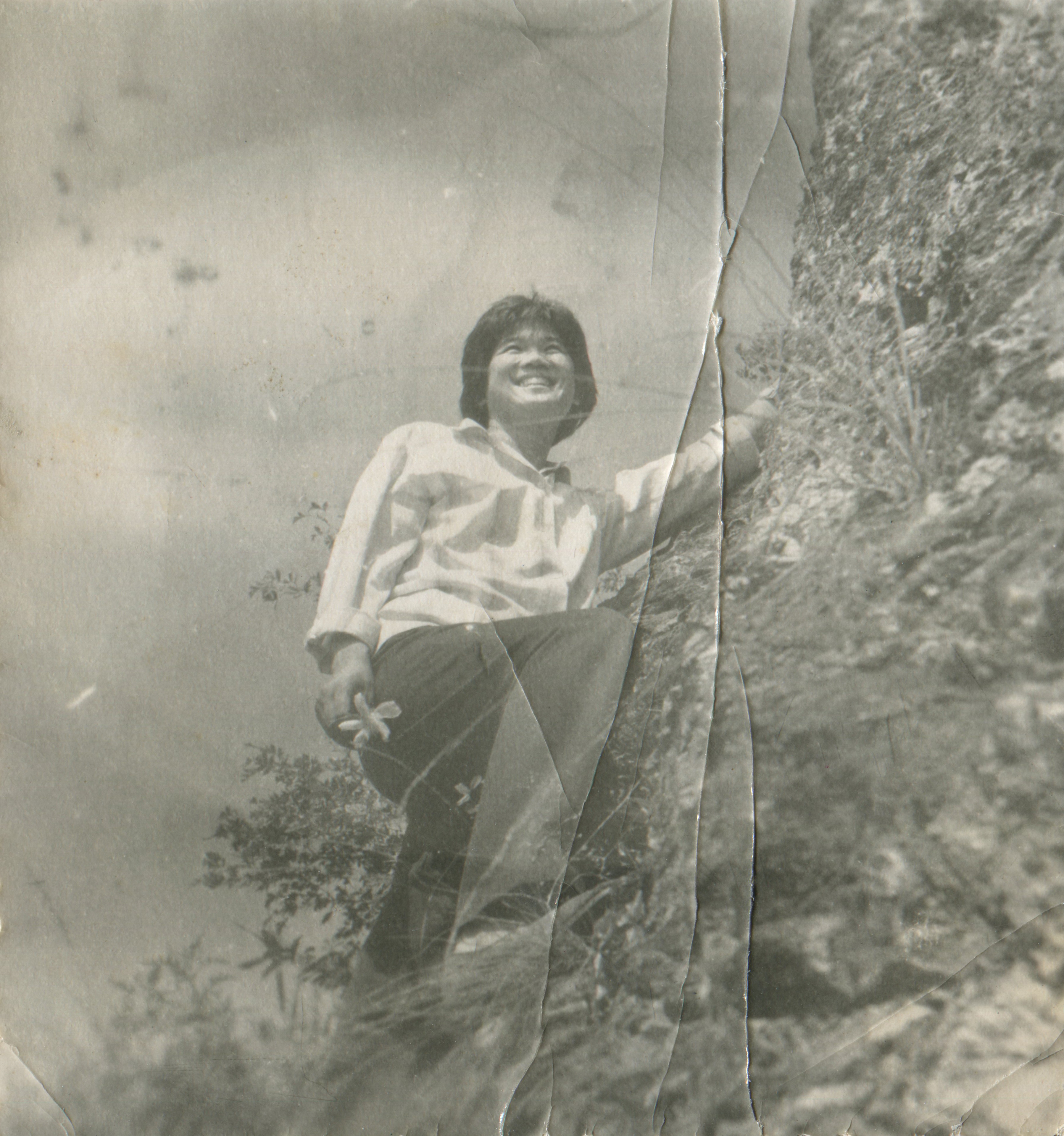 Person som smiler mens han klatrer en steinete skråning, iført en uformell skjorte og mørke bukser. Bildet har en vintage følelse med synlige bretter