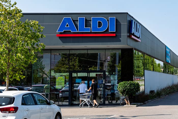 People entering an ALDI store with a shopping cart in the parking area. A white car is parked in the foreground