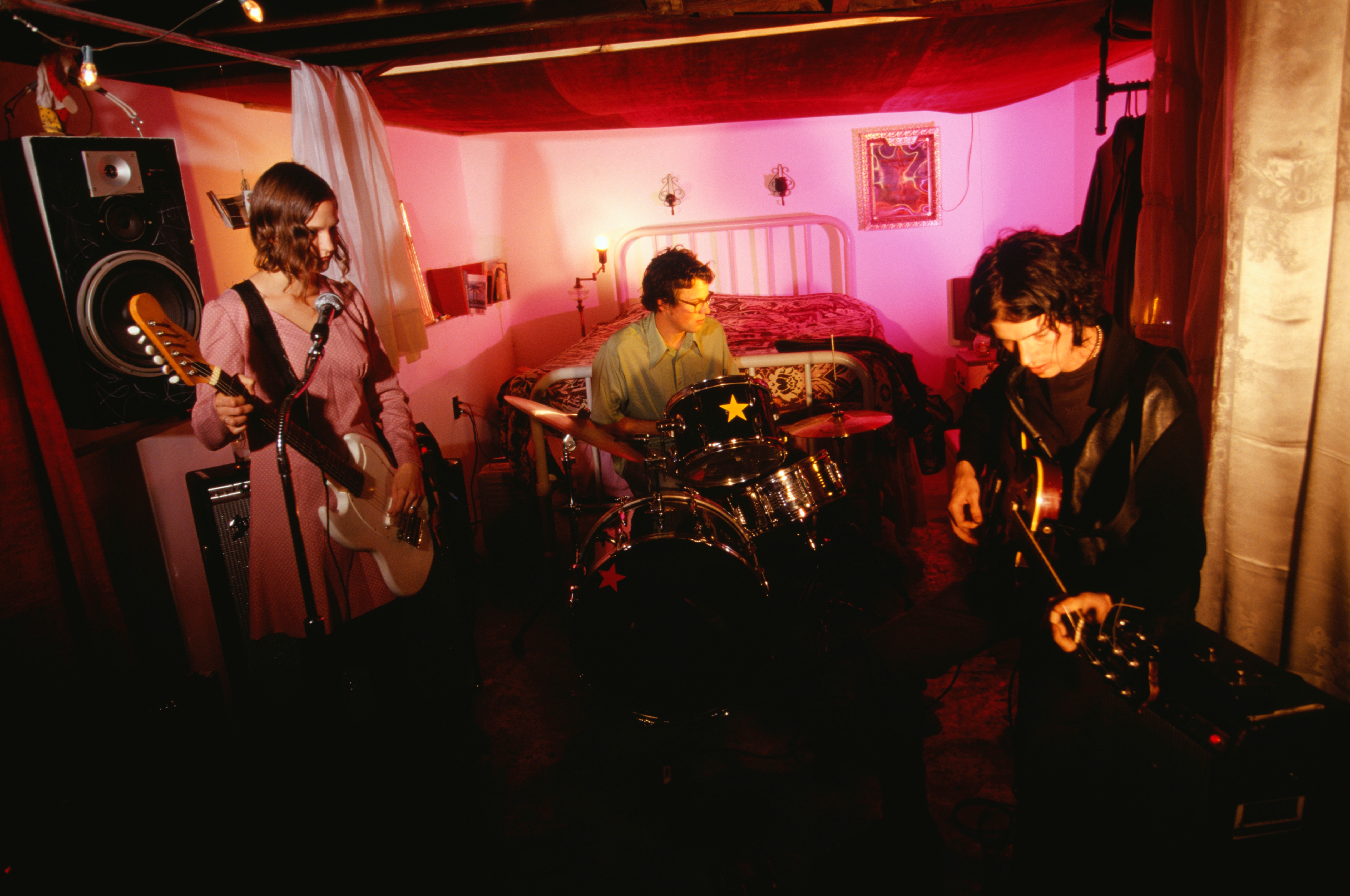 Tre musikere spiller instrumenter i et koselig rom: en på gitar, en på bass og en på trommer, og skaper en intim forestillingssetting