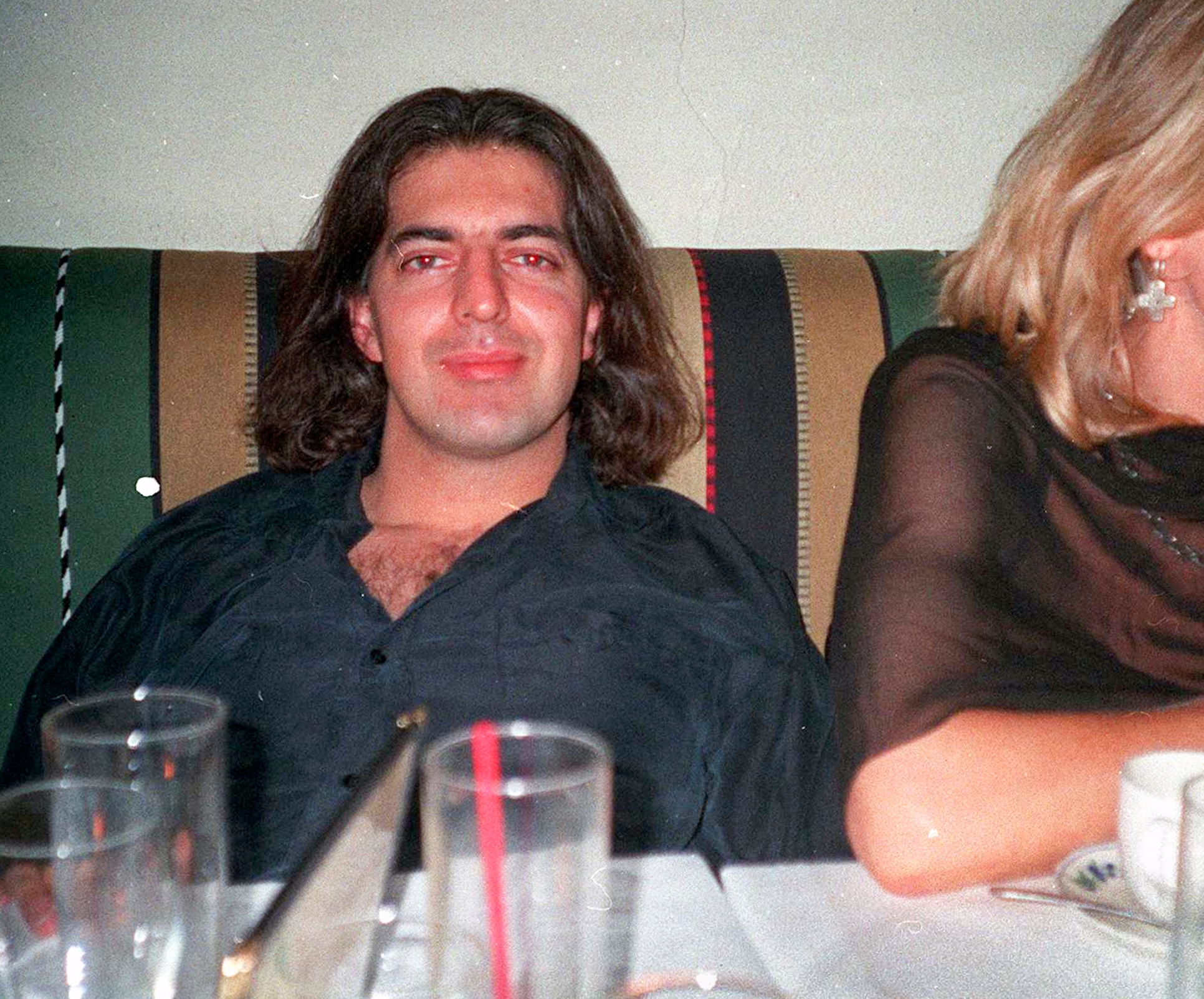 En person med skulderlangt hår sitter ved et spisebord og smiler. De har på seg en uformell skjorte med åpen krage