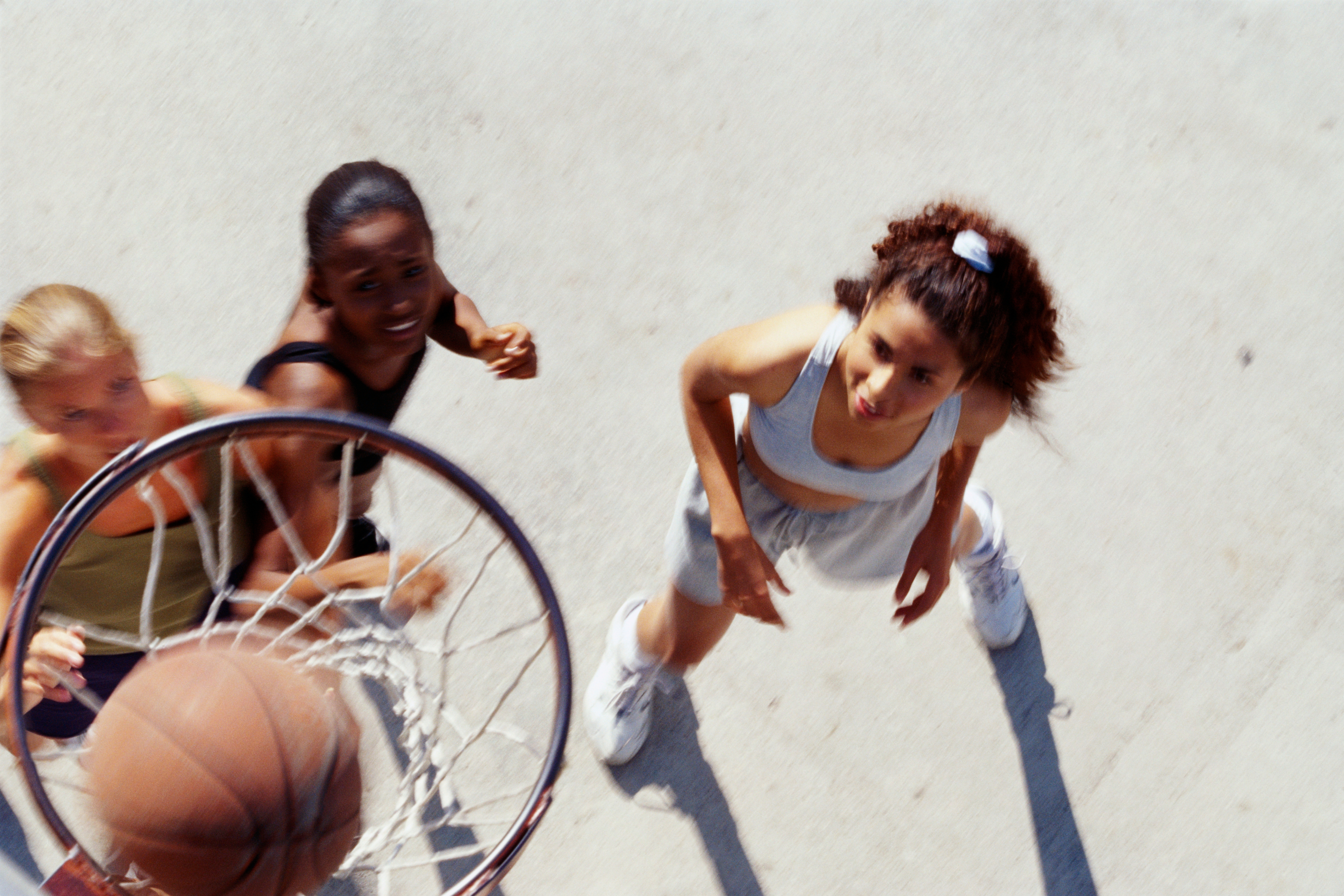 Tre personer som spiller basketball på en utendørsbane; fokus på ballen som går gjennom bøylen