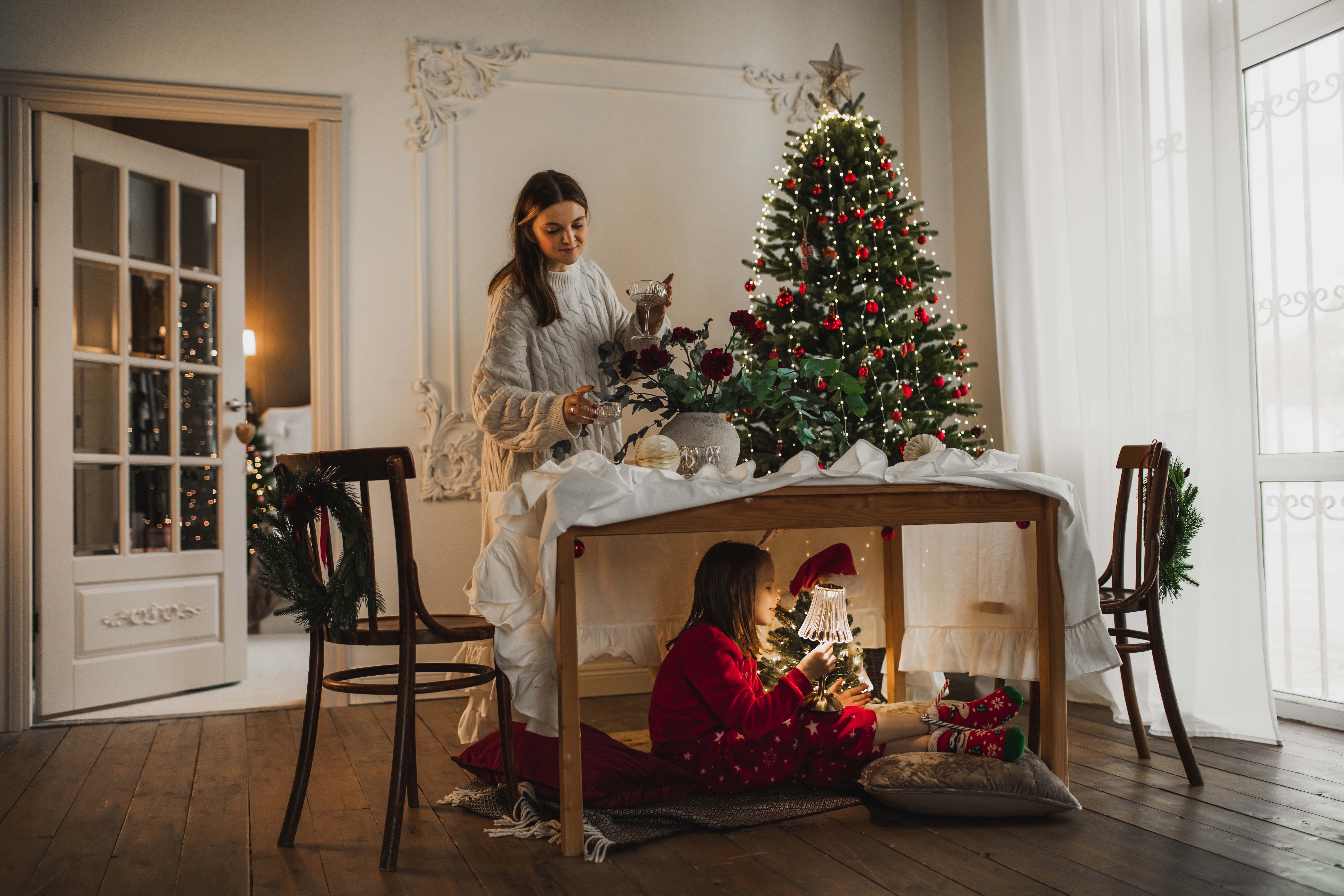 En kvinne dekorerer et festlig bord nær et juletre; et barn i rødt antrekk leser under, og skaper en koselig feriescene
