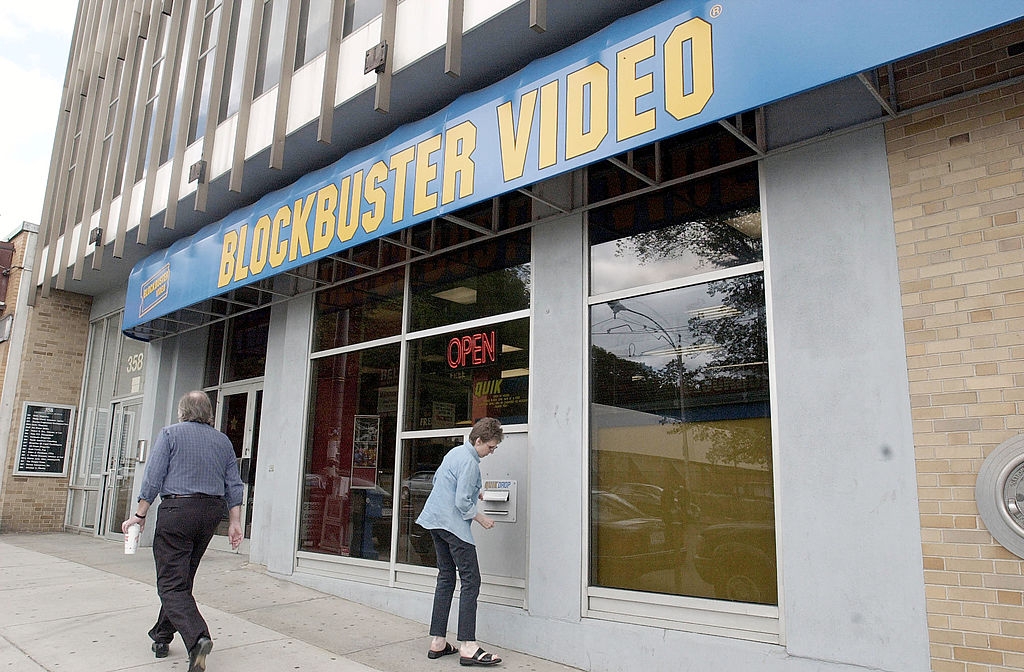 People walking by a Blockbuster Video store, with 1  idiosyncratic   utilizing an outdoor kiosk