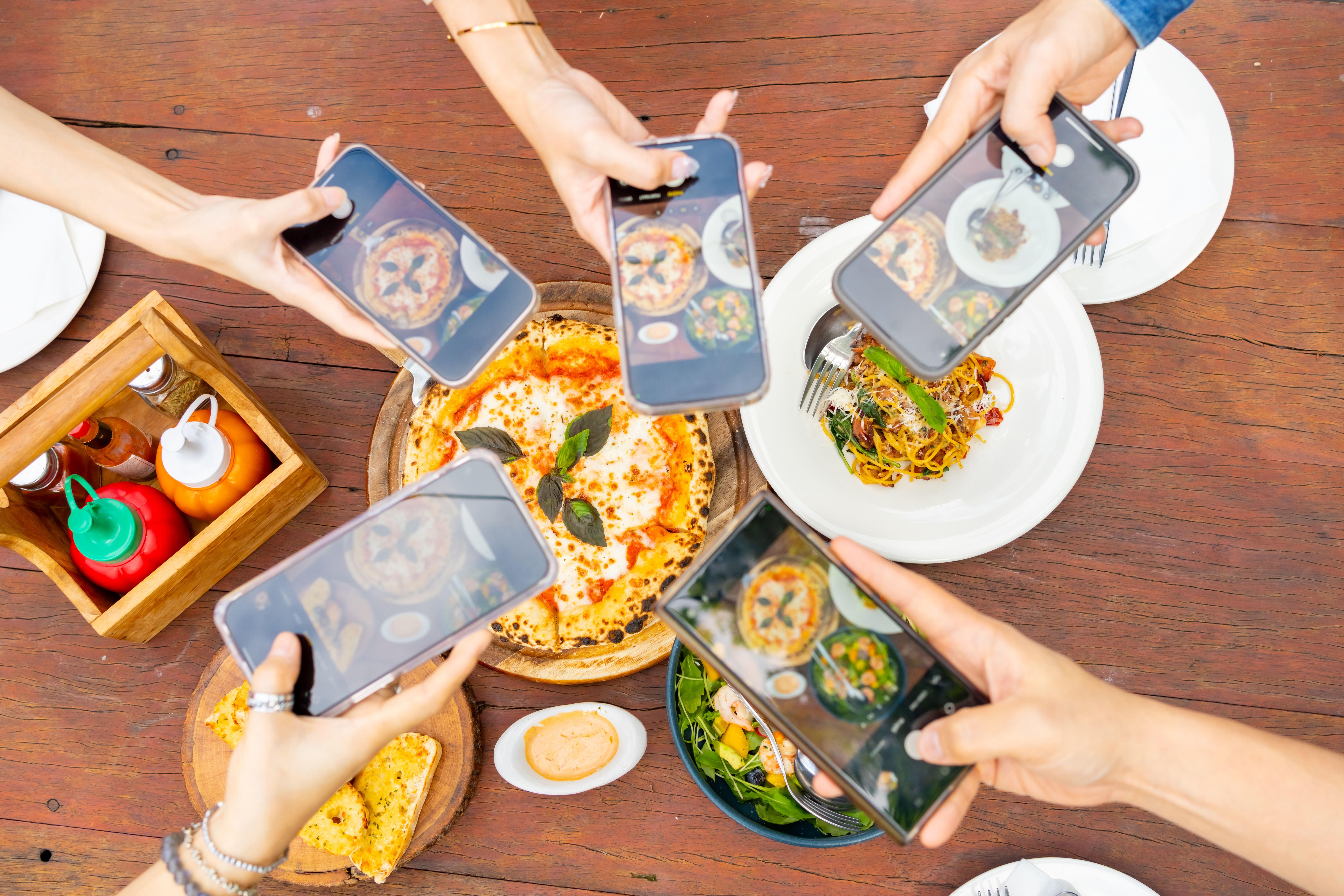 People taking photos of a pizza and pasta on a wooden table with smartphones, capturing the meal from different angles