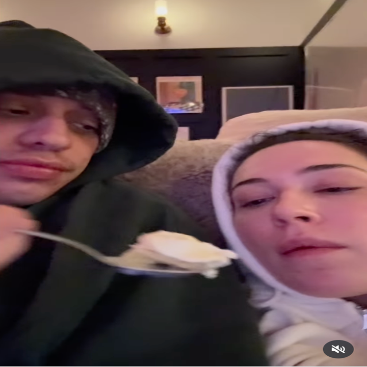 Two people in hoodies are relaxing at home. One offers the other a spoon of food. Casual and cozy setting