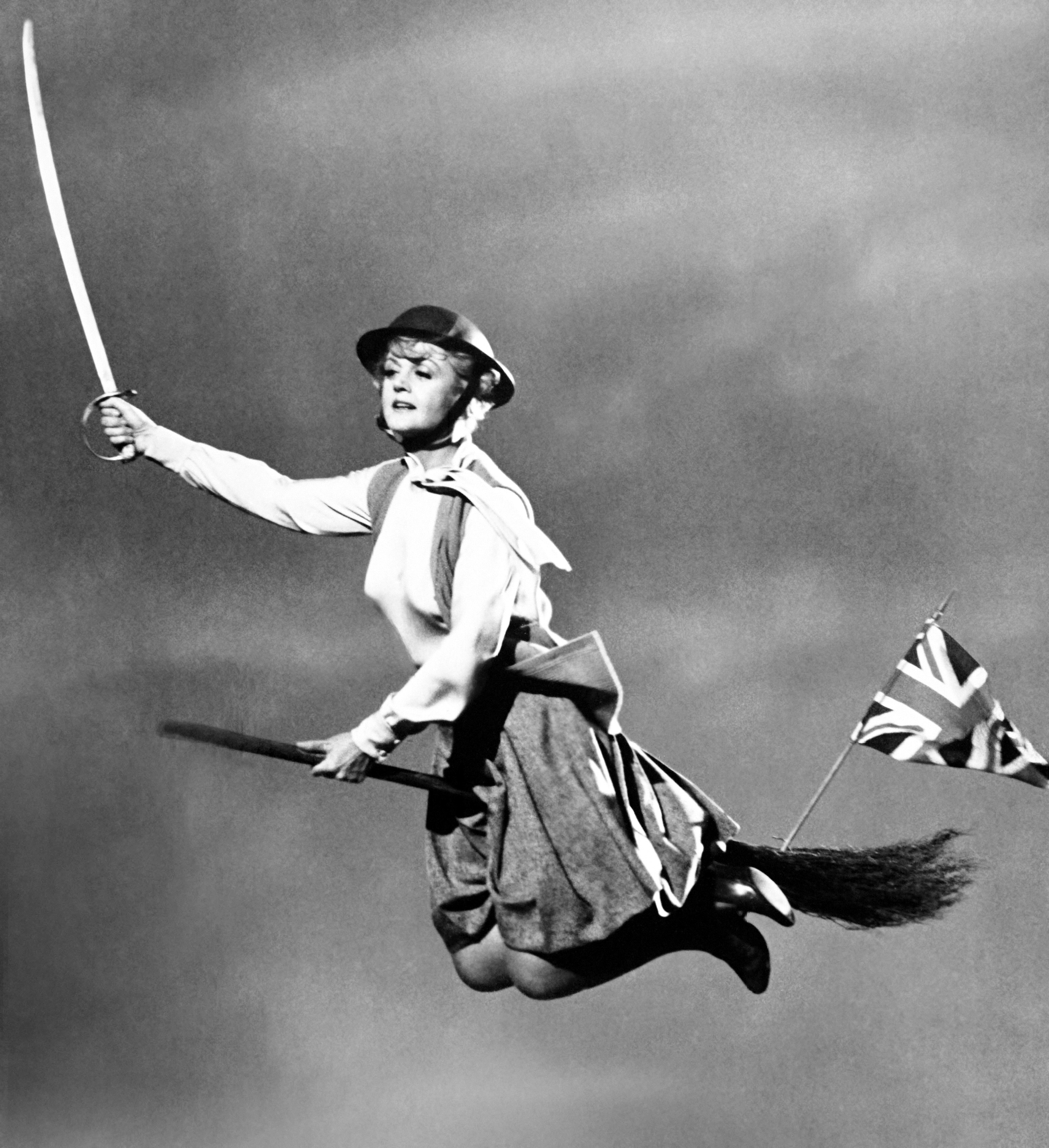 Angela Lansbury successful  vintage attire, helmet, and boots flies connected  a broomstick holding a sword and British flag, resembling a whimsical, magical scene