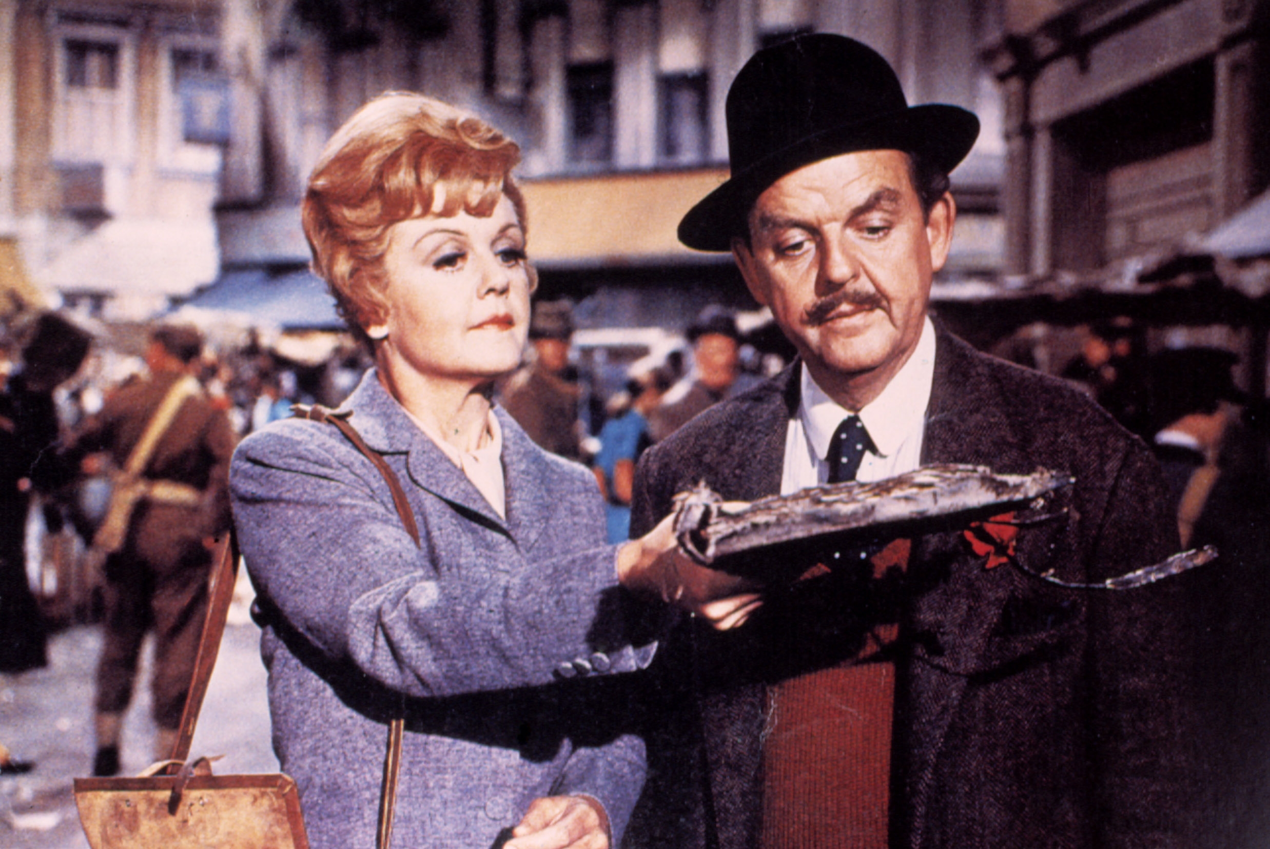 Angela Lansbury and David Tomlinson successful  vintage attire are lasting  successful  a thoroughfare  scene. The pistillate   holds a tray covered with an antithetic   item. They look  focused