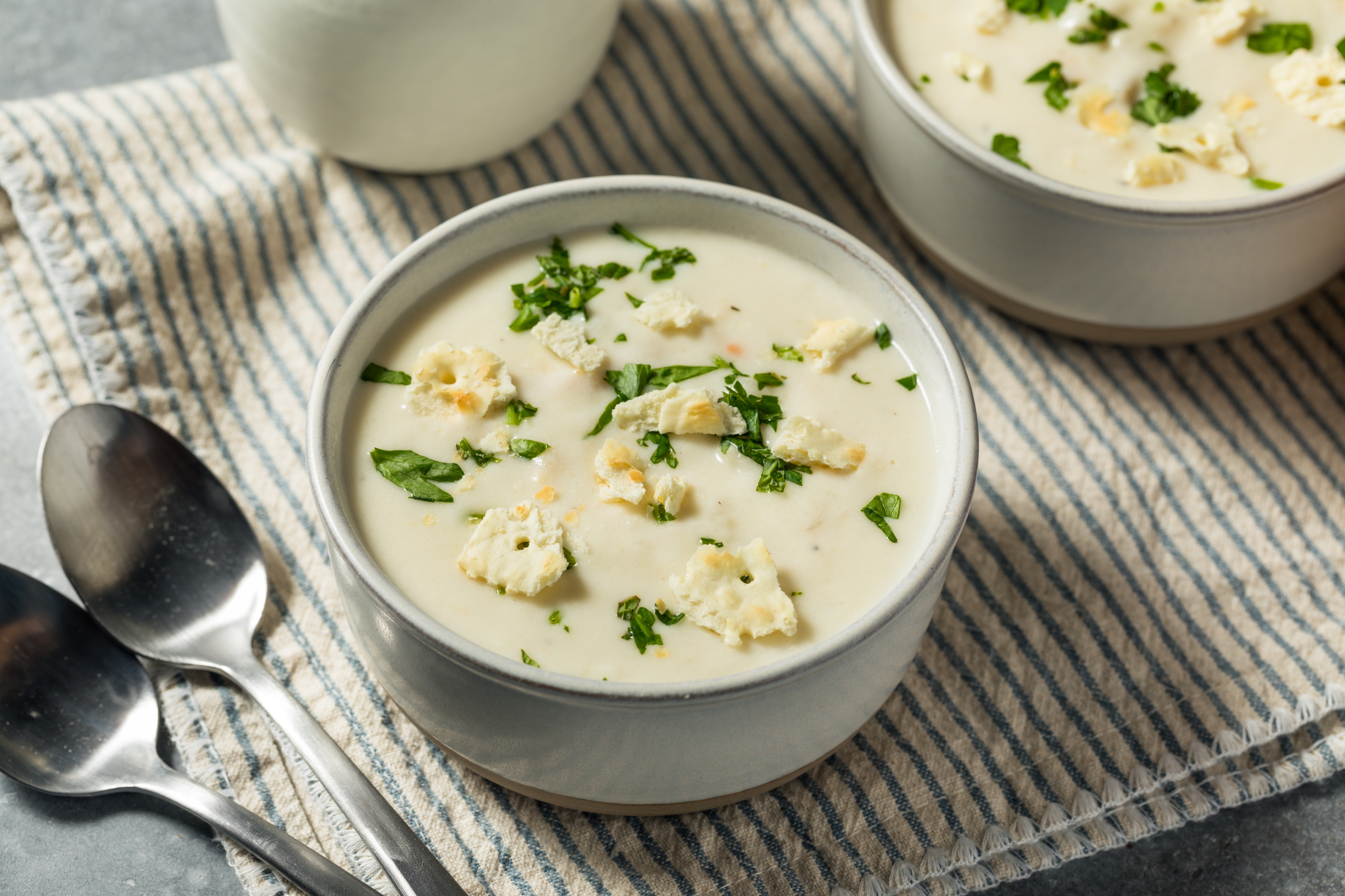Bowls of creamy soup topped with crumbled cheese and fresh herbs, placed on a striped cloth, accompanied by two spoons