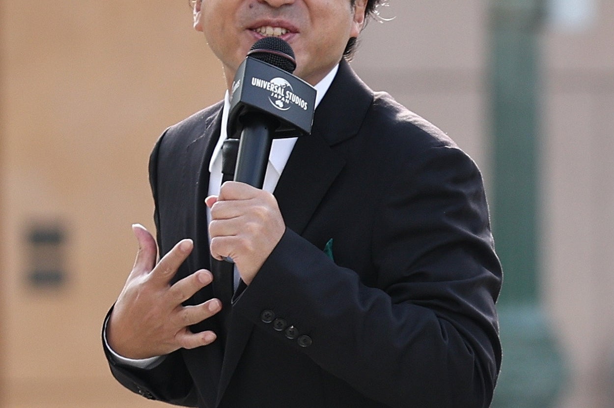 マイクを持ちながらスーツ姿で話す男性。髪はふんわりしており、カメラに向かってジェスチャーをしています。