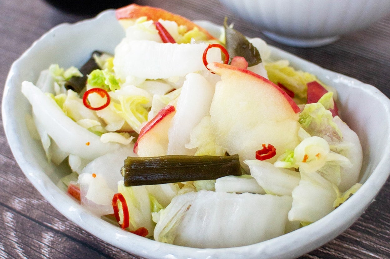 キムチや唐辛子が添えられた白菜とリンゴの漬物が白い陶器の器に入っており、横にご飯と醤油が置かれています。