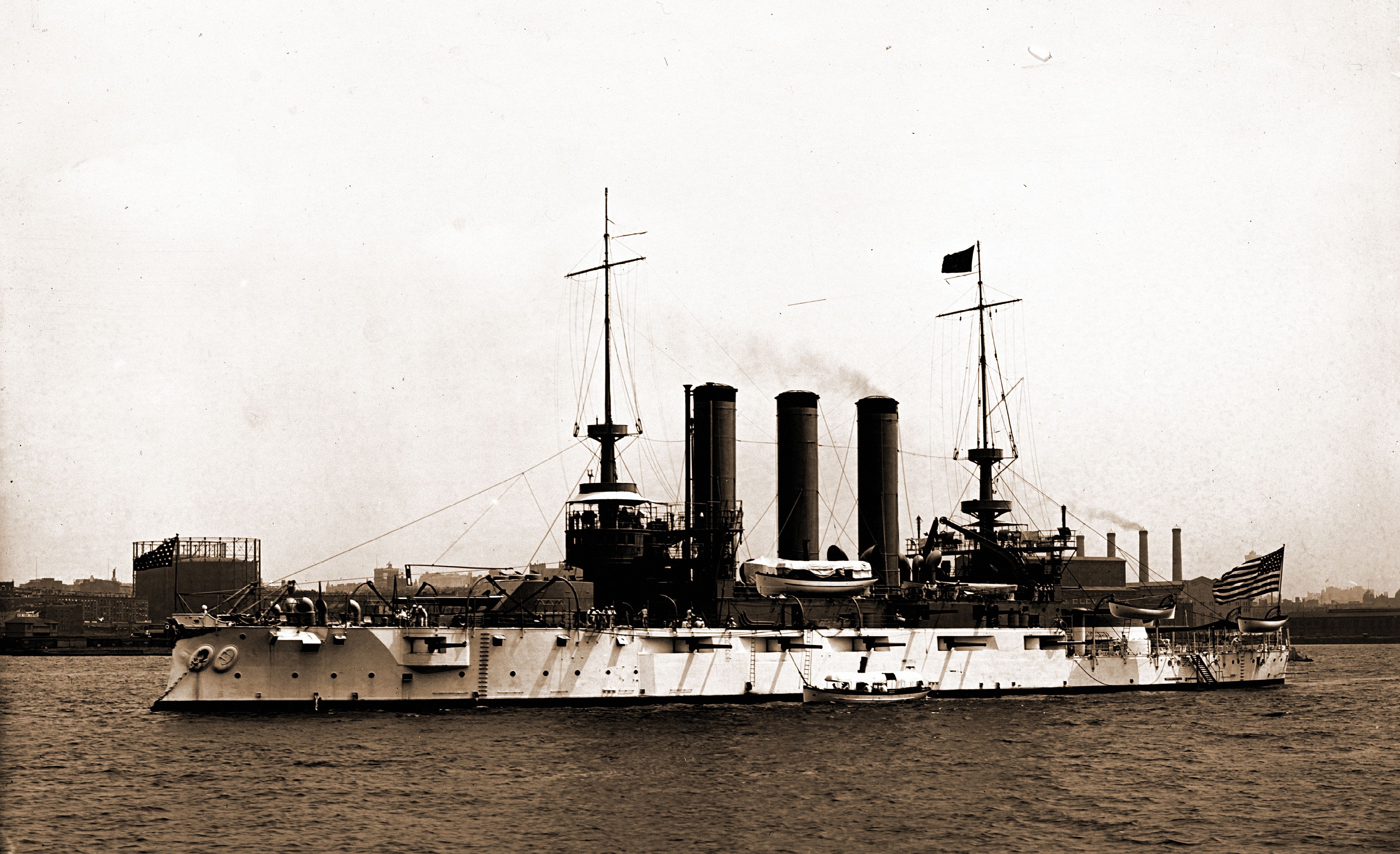 Historical naval battleship with 3  ample  smokestacks and respective  masts connected  the water, silhouetted against an concern   cityscape