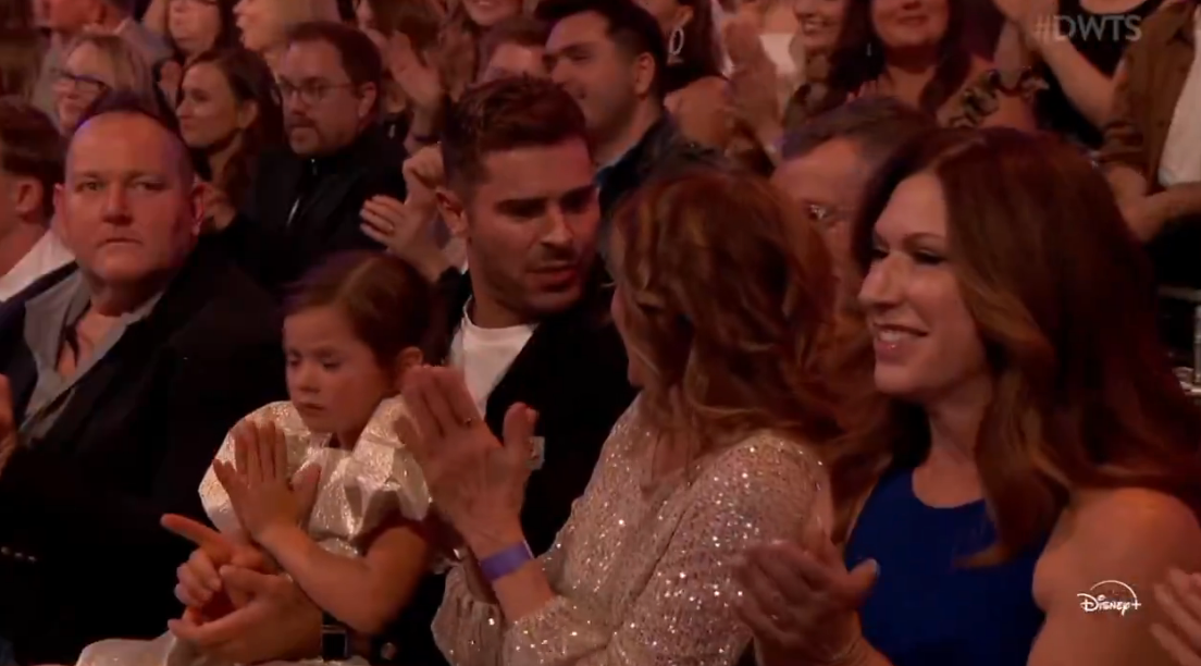 Audience astatine  an lawsuit   applauding; a young miss  sits adjacent  to an adult, surrounded by others clapping