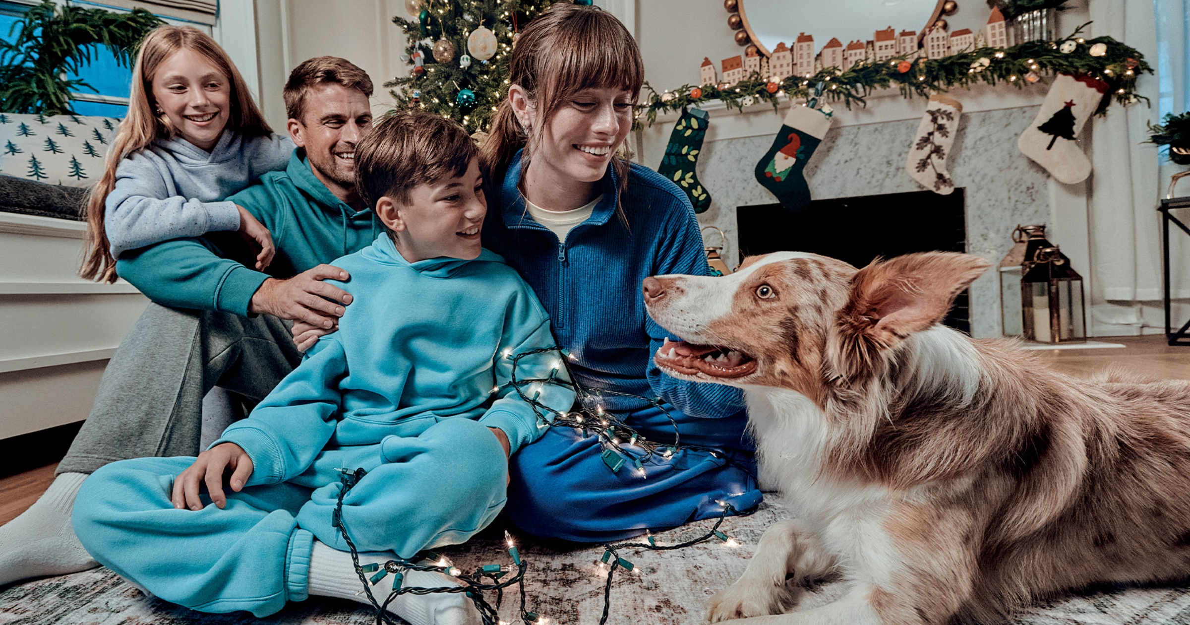 Family gathered in a cozy living room during a holiday, seated near a decorated fireplace with stockings, and warmly interacting with their dog