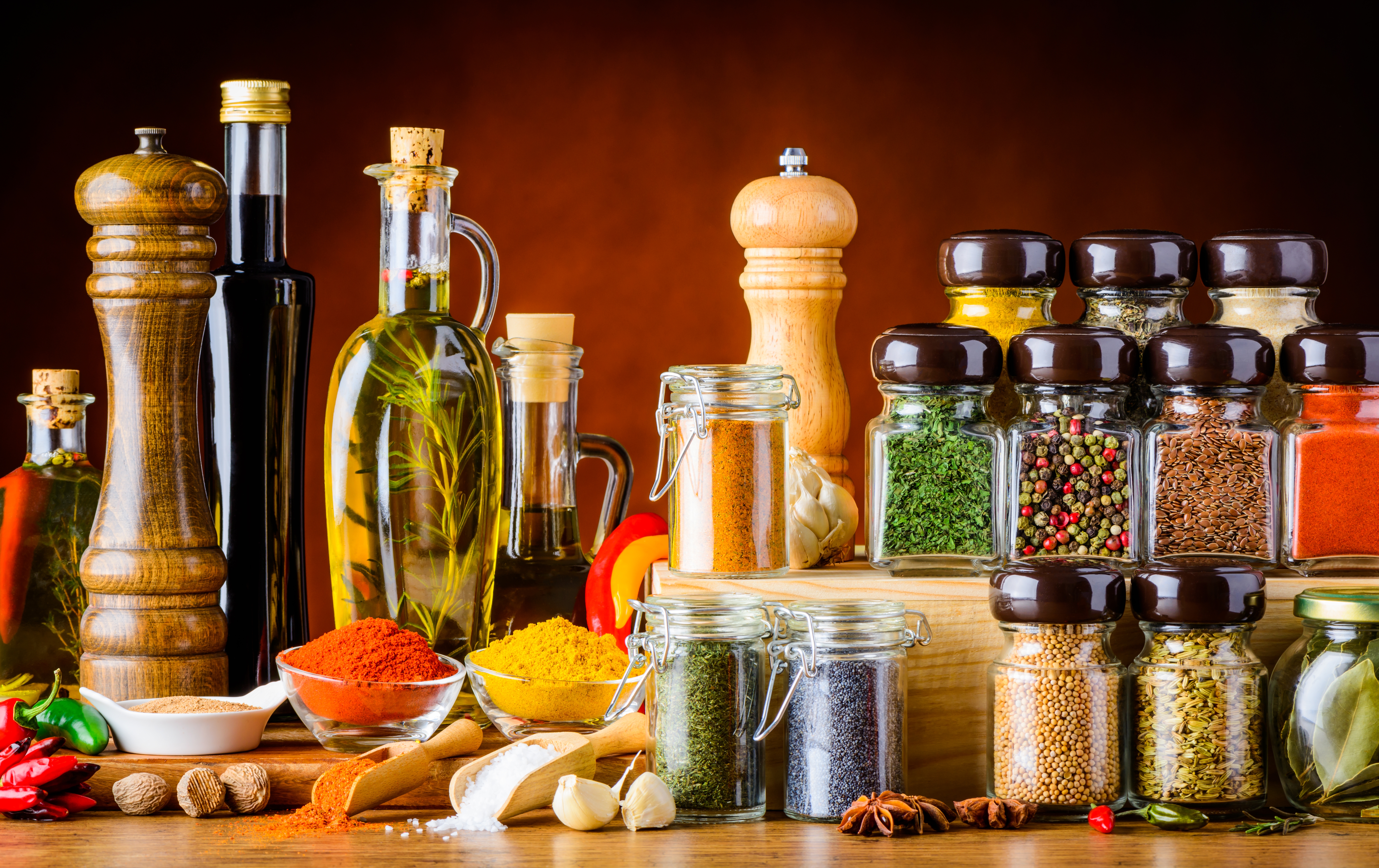 Assorted spices and herbs in jars and bottles are displayed on a wooden surface, including pepper mills, oils, and various ground and whole spices