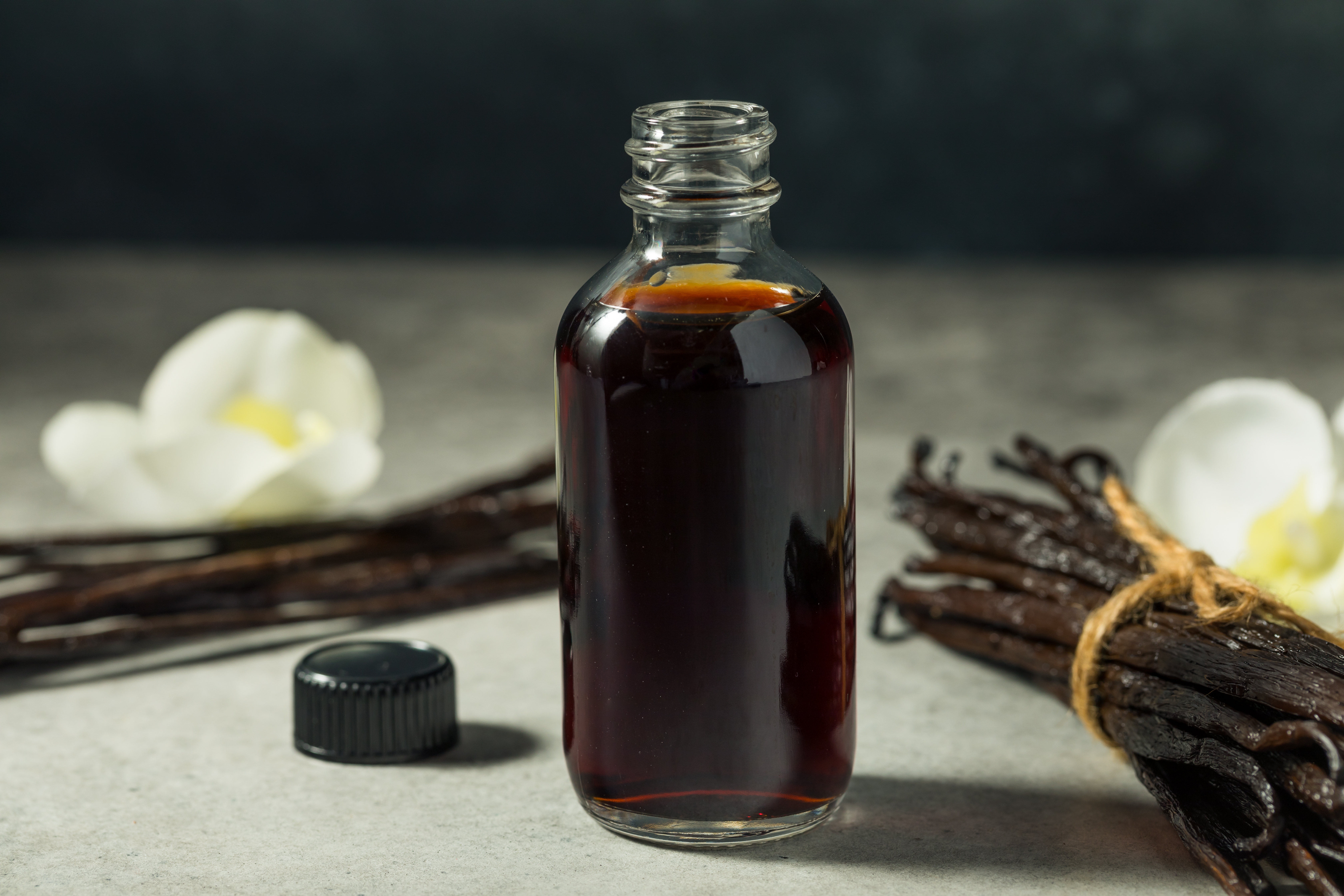 A bottle of vanilla extract next to vanilla beans and white flowers on a gray surface