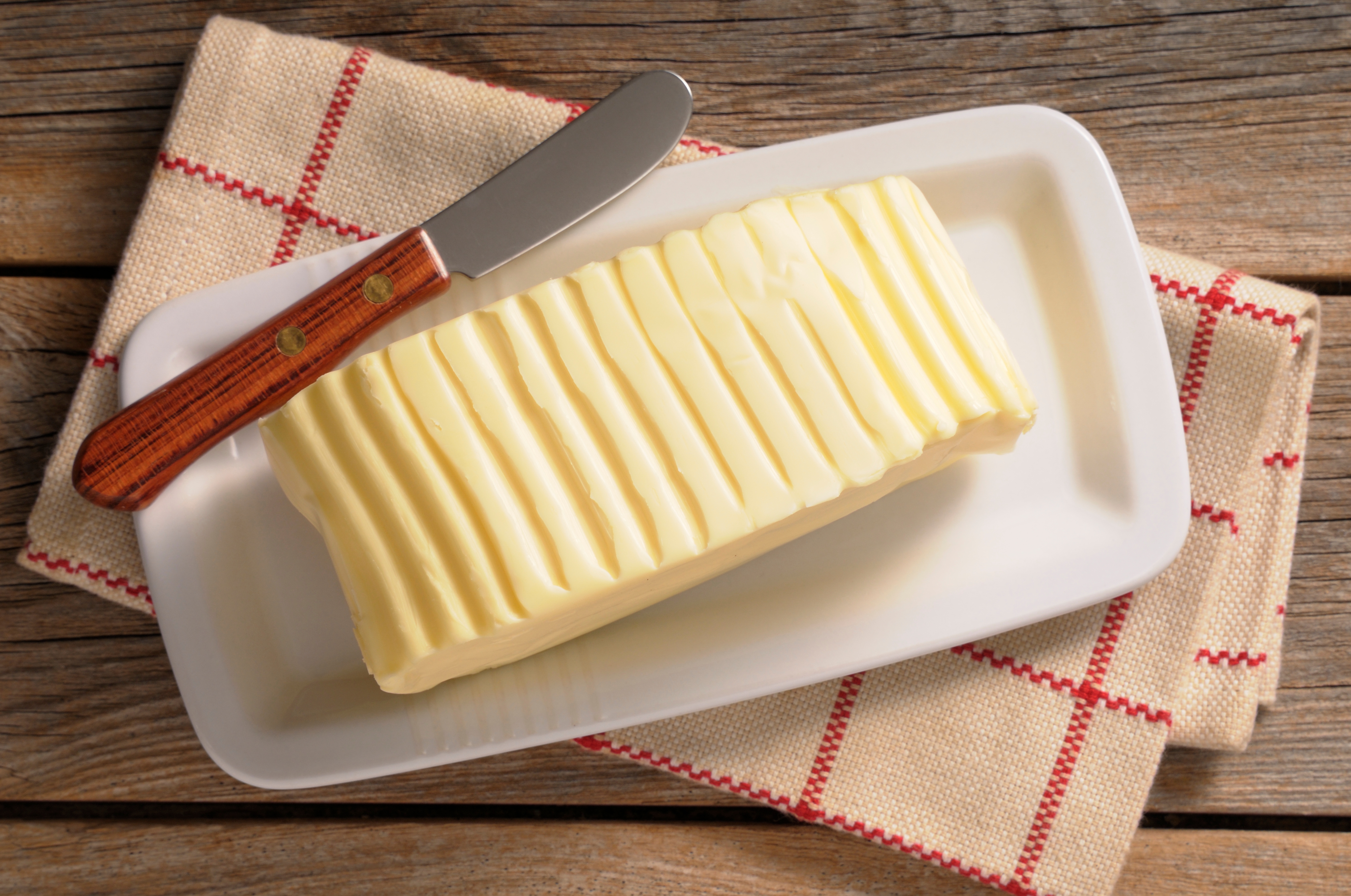 Sliced stick of butter on a white dish with a wooden-handled butter knife, set on a checkered cloth atop a wooden surface