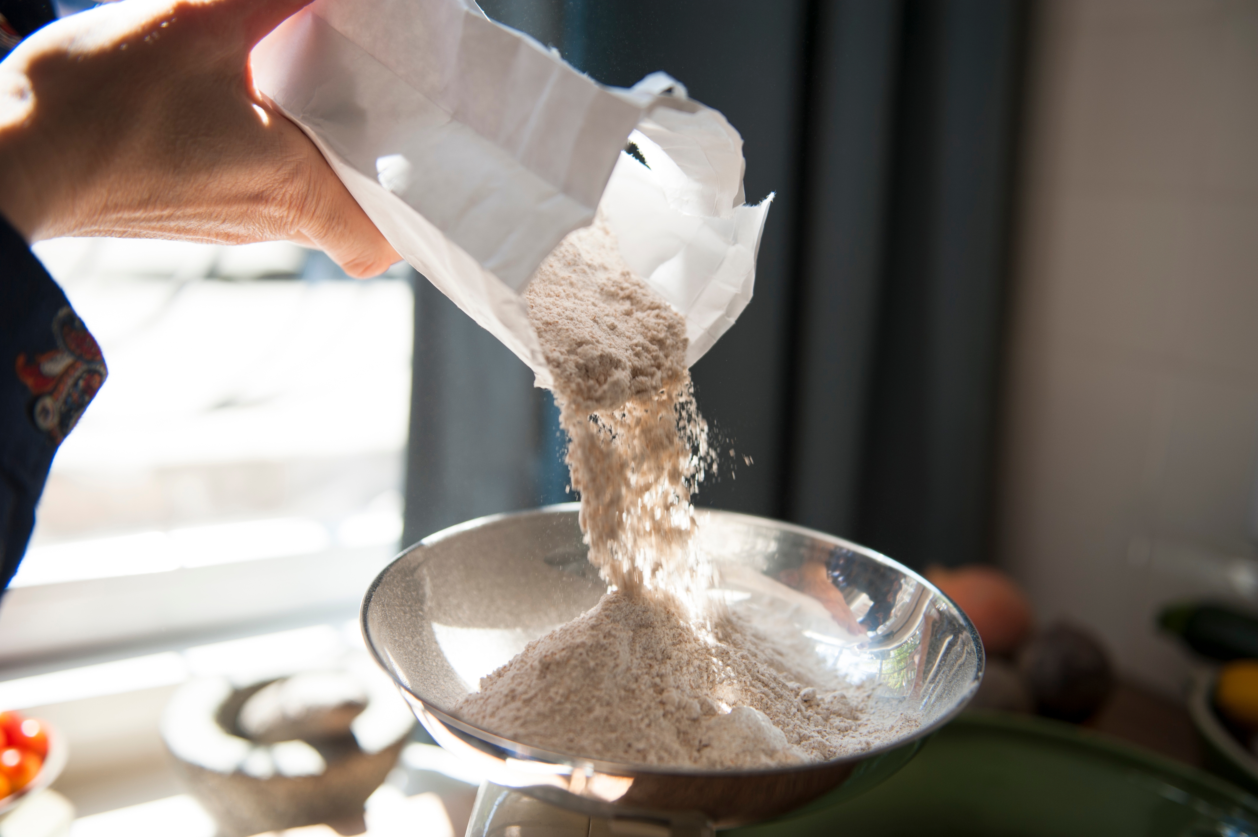 Hand pours flour from a bag into a kitchen scale bowl, sunlight in the background