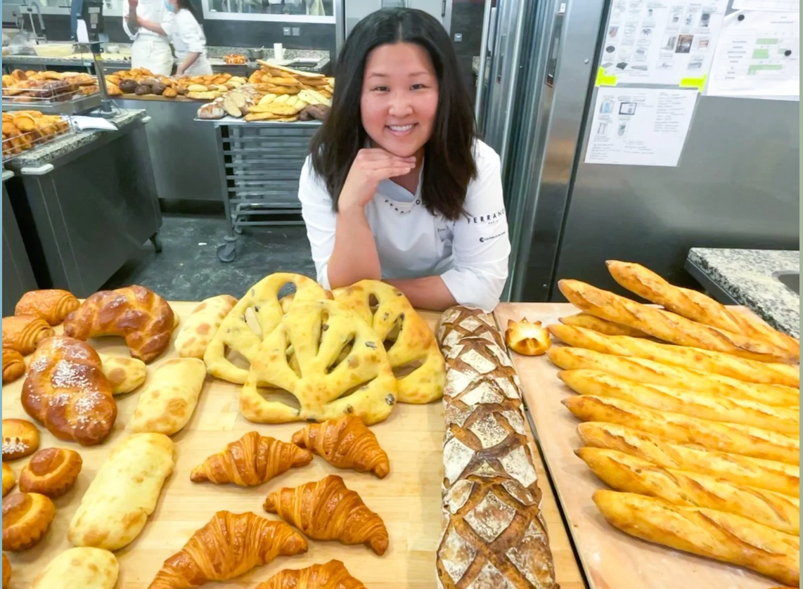 Jen Kim in a bakery kitchen smiling behind a large assortment of artisanal breads and pastries on display