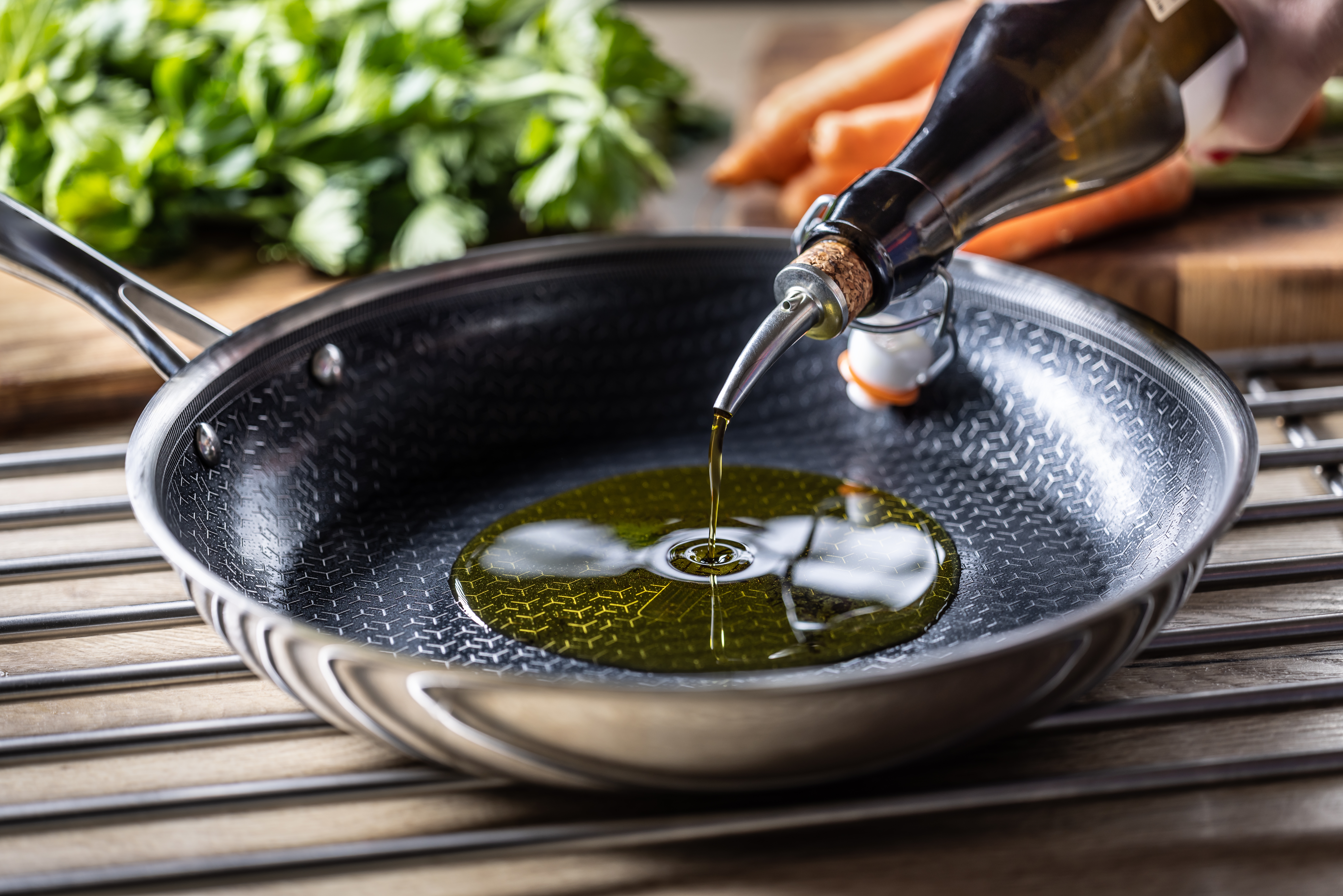Oil being poured into a frying pan on a stove, with fresh herbs and carrots in the background