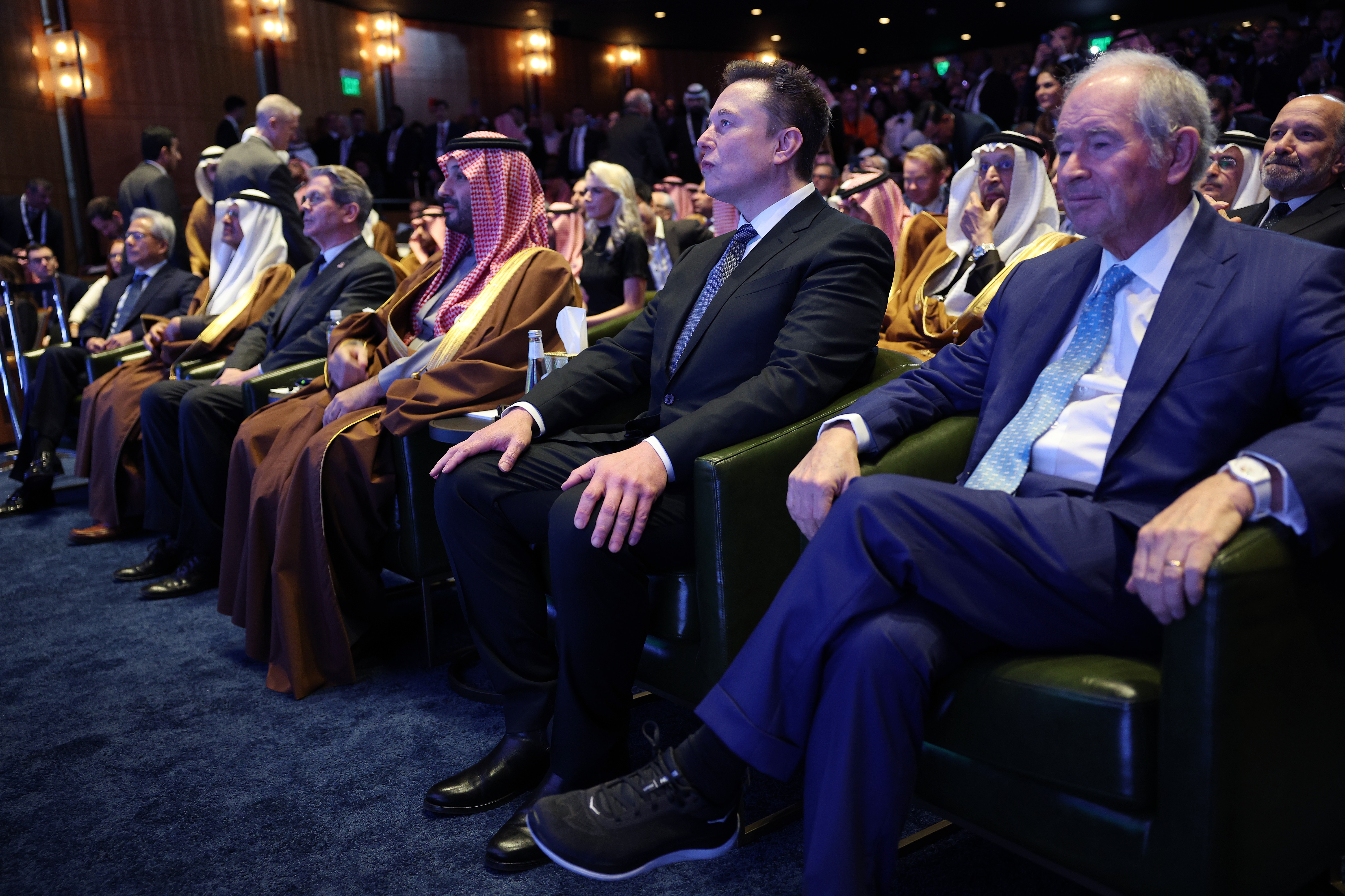 A group of formally dressed people, including a man in a suit and sneakers, sit in an audience at a formal event