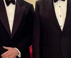 Four individuals in formal tuxedos pose in a grand setting with ornate decor, including a staircase and a prominent painting in the background