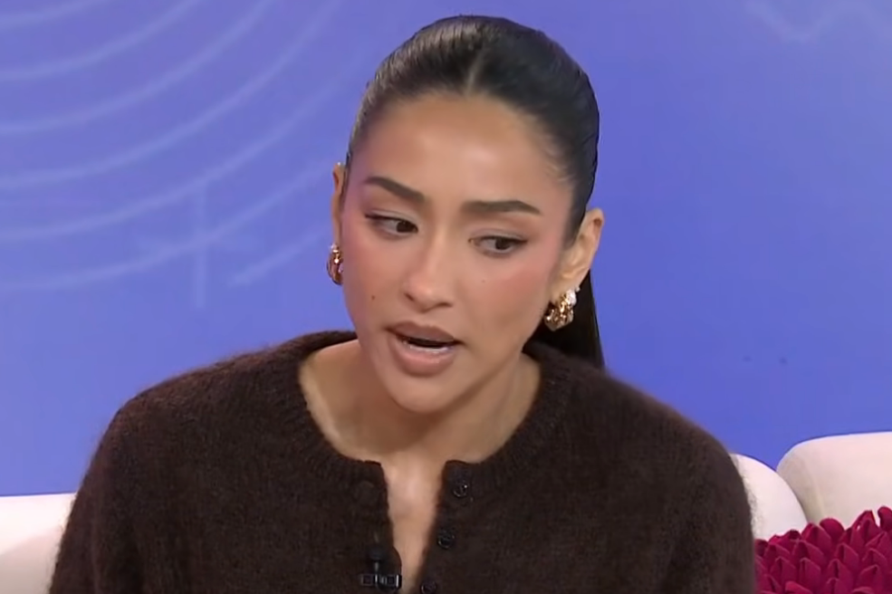 Shay Mitchell with long hair speaking on a talk show, wearing a textured cardigan