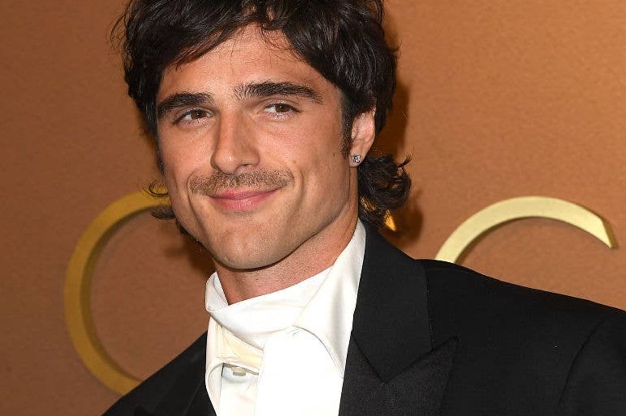 Man in formal attire with a tuxedo jacket and white shirt, smiling at a celebrity event backdrop