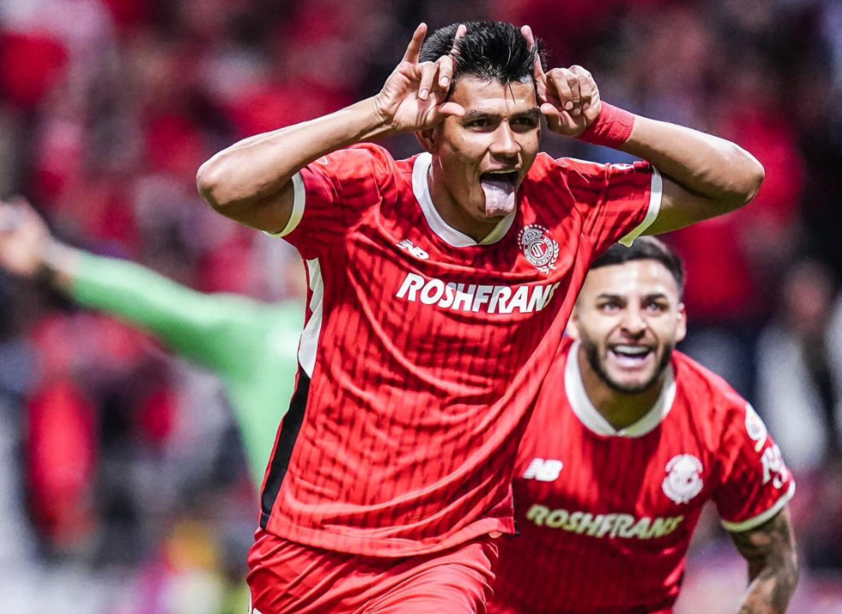 Soccer players celebrate on the field, one making a playful gesture with hands on head and tongue out