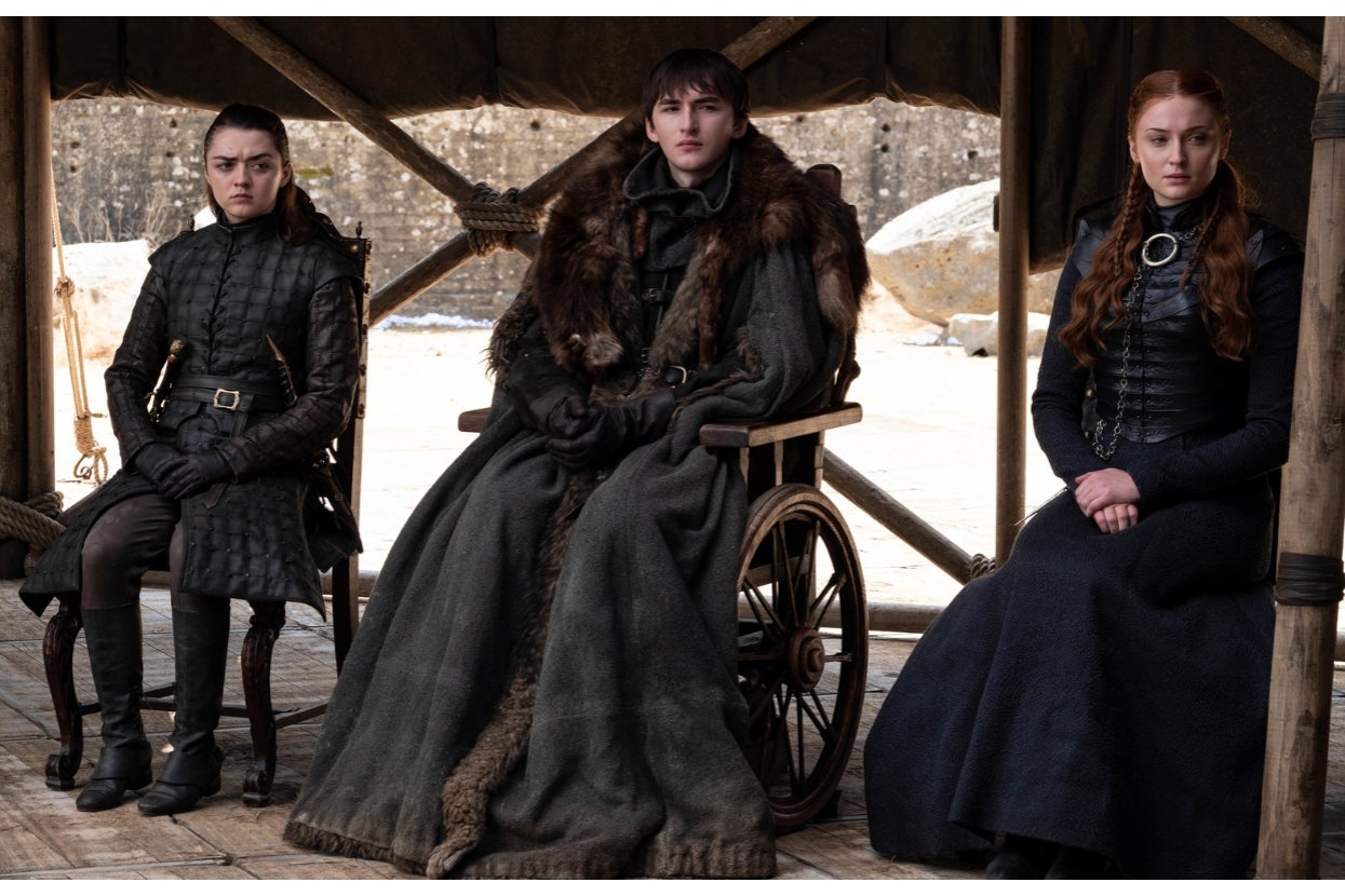 Three characters from a medieval fantasy scene sit closely, one in a wheelchair, all wearing dark, detailed attire with leather and fur accents