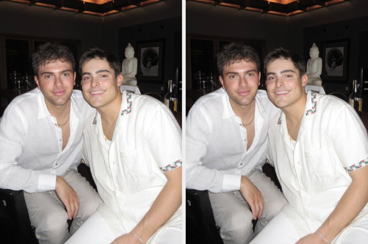 Two people pose closely together indoors, both wearing casual open white shirts, smiling. Background features art and figures