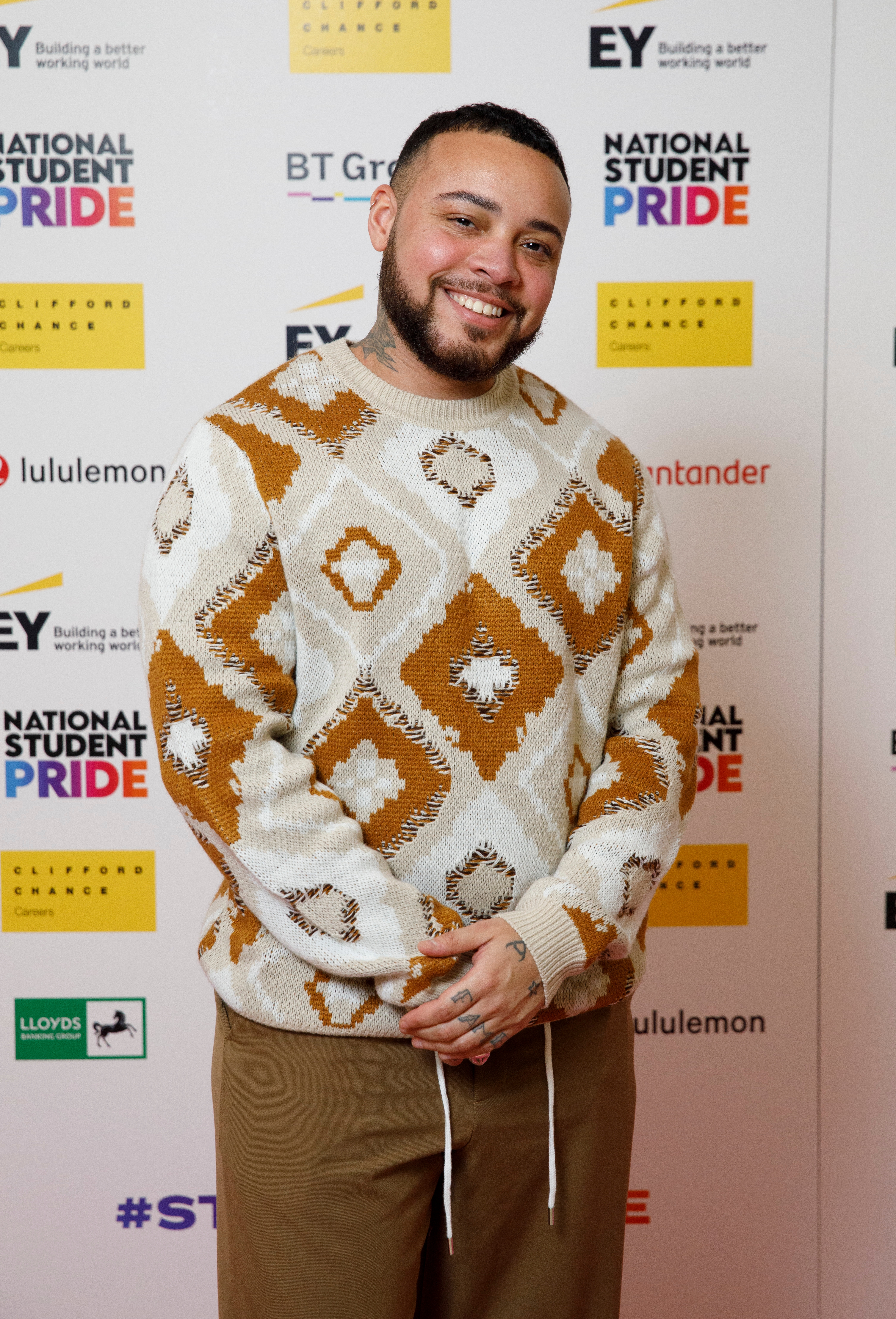 Person som smiler på den røde løperen på National Student Pride-arrangement, iført en mønstret genser og brune bukser