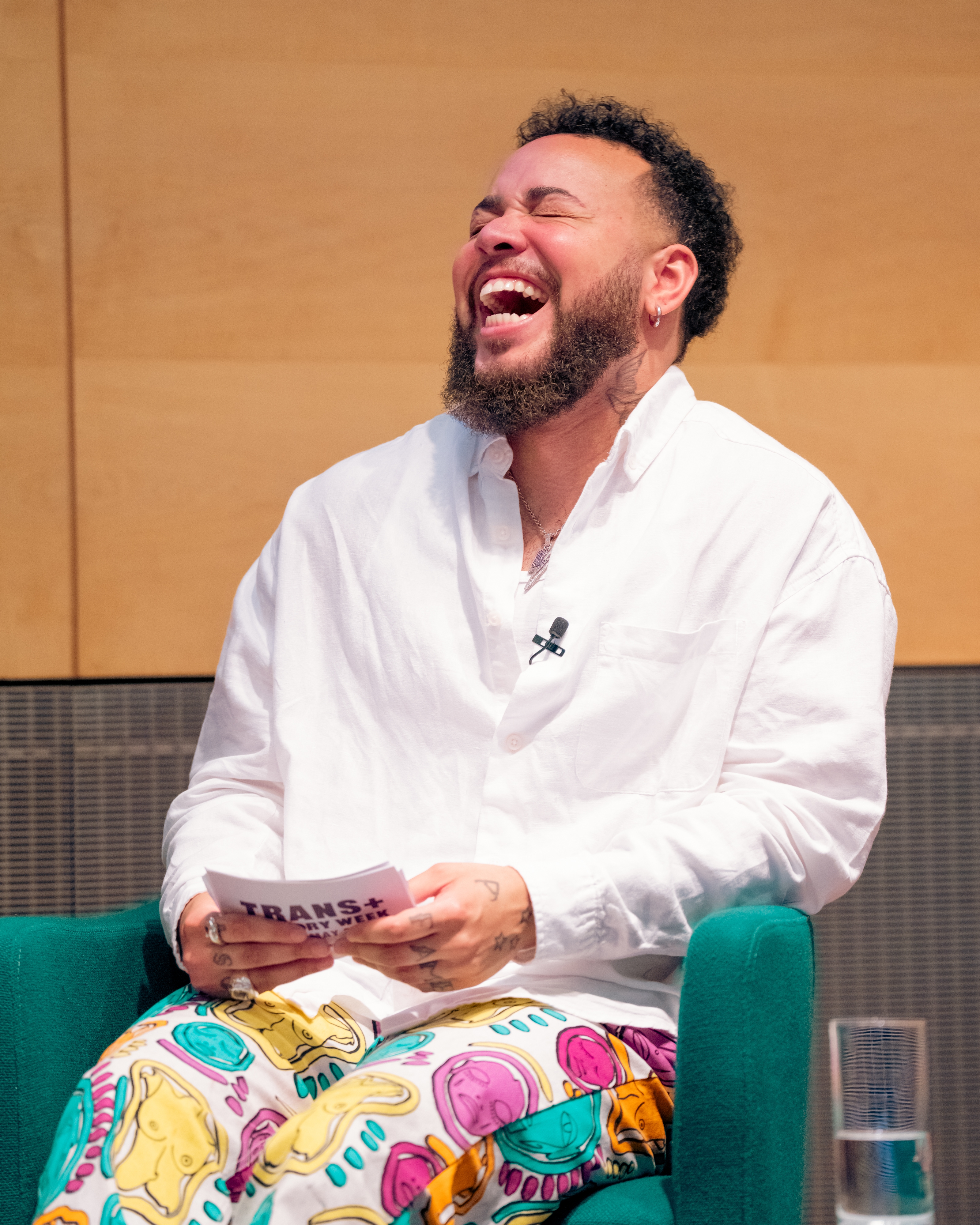 Person som ler glad, iført en hvit skjorte og fargerike bukser med abstrakte mønstre, holder et kort under en foredrag eller paneldiskusjon