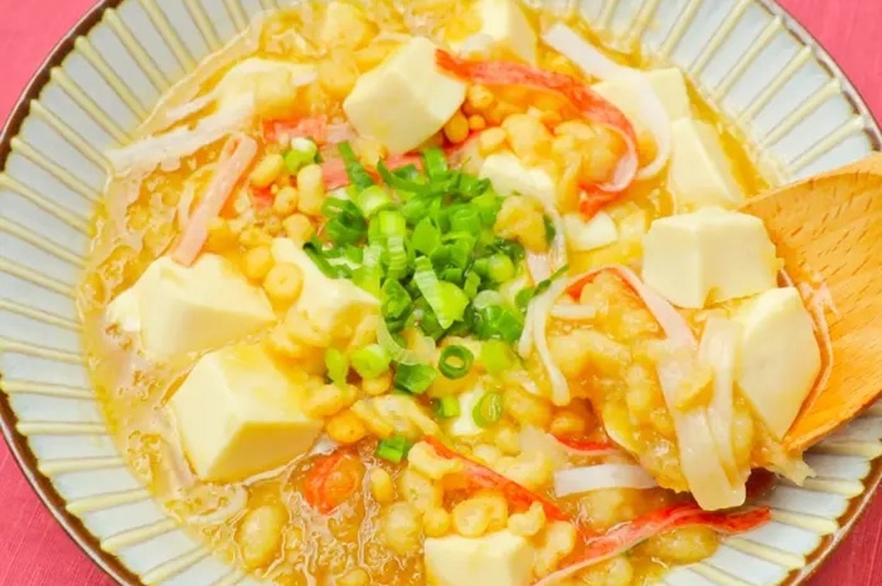 豆腐とカニカマが乗ったあんかけ料理。ネギがトッピングされ、木のスプーンが添えられている。