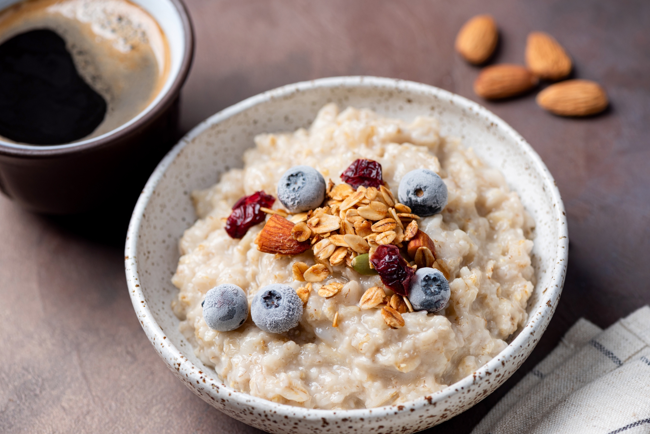 Bowl of oatmeal topped with blueberries, almonds, cranberries, and granola adjacent  to a cupful  of coffee