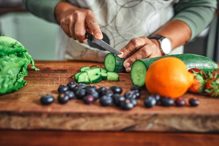 Person slicing cucumber connected  a cutting committee  with lettuce, blueberries, an orange, and a strawberry nearby