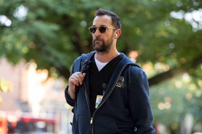 A idiosyncratic   wearing sunglasses, a hoodie, and wireless earbuds walks outside, carrying a backpack