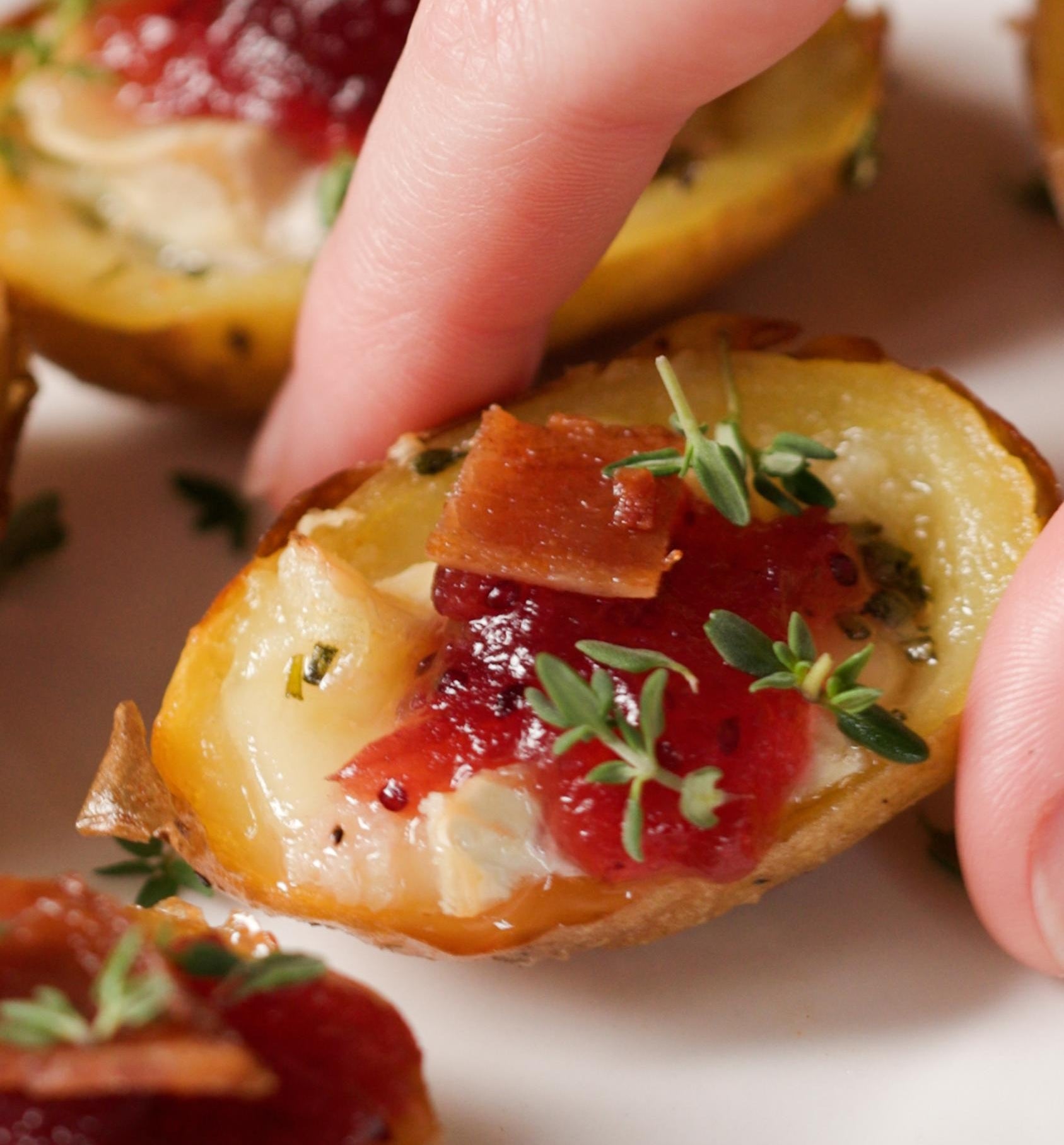 Hand reaching for mini potato skin topped with cheese, bacon, cranberry sauce, and herbs on a plate, perfect for a savory appetizer