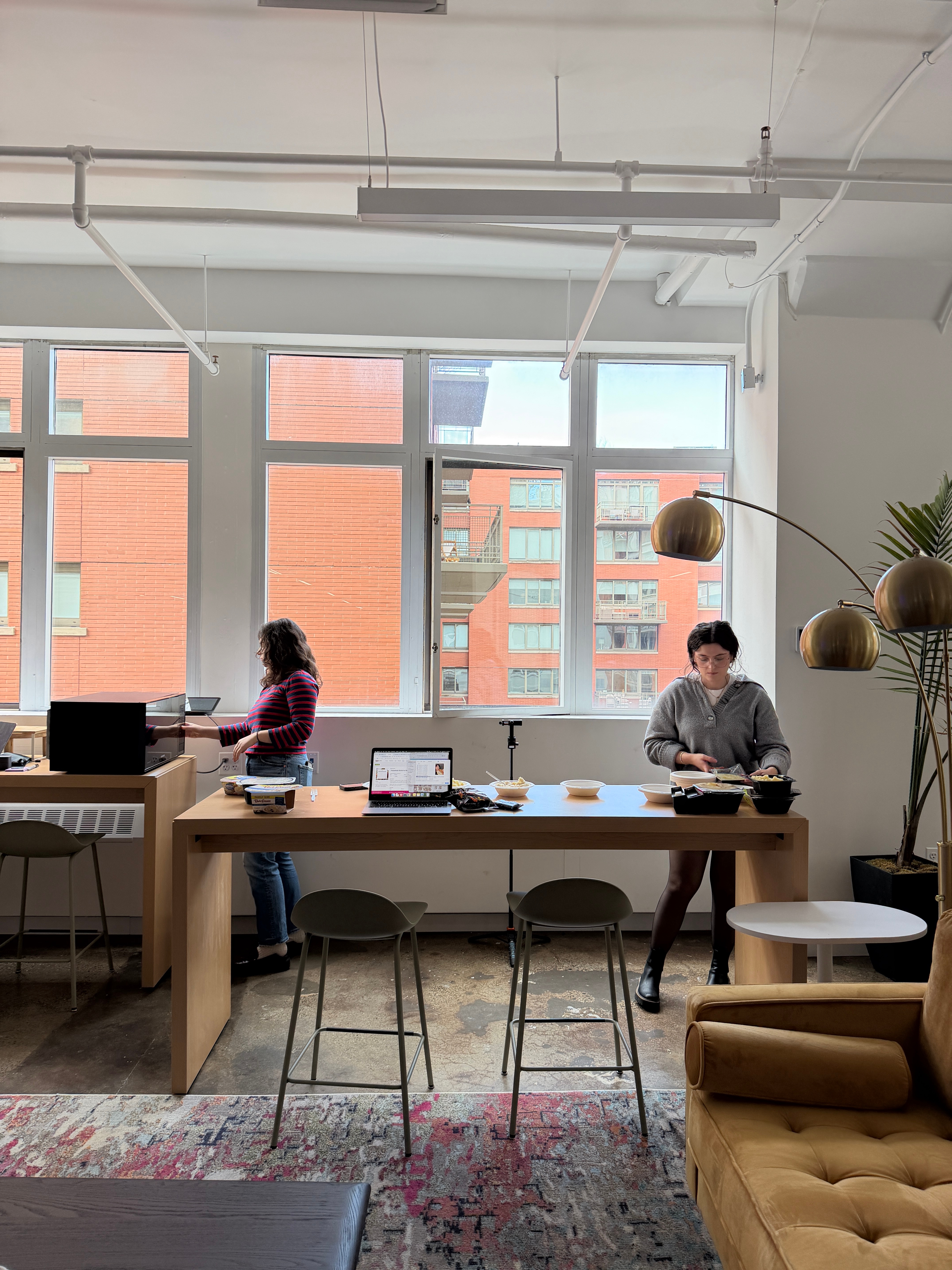 Two people work at a shared table with laptops and equipment in a bright office with large windows
