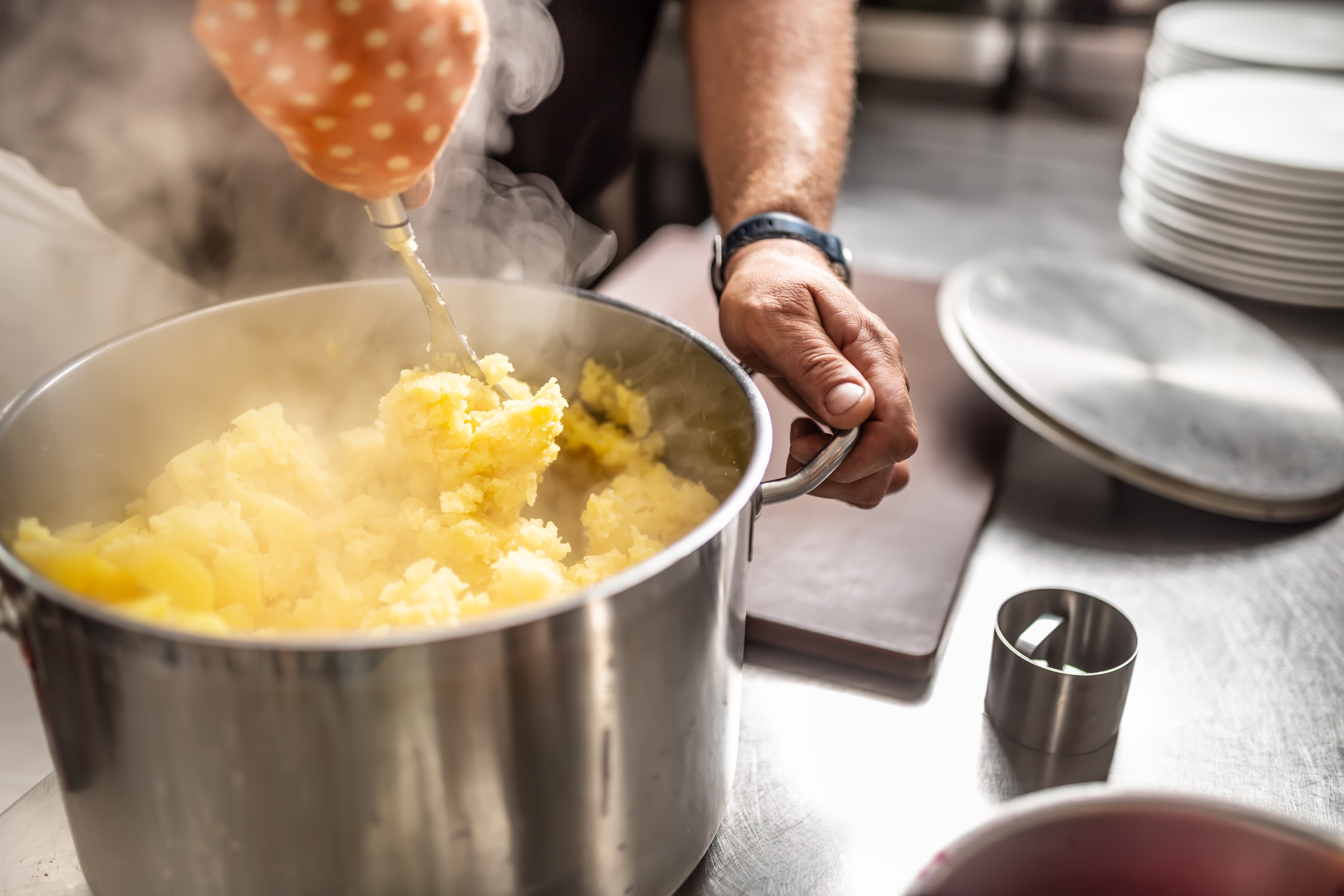 Person mashing potatoes in a large pot on a kitchen counter, with steaming rising, suggesting they&#x27;re freshly cooked