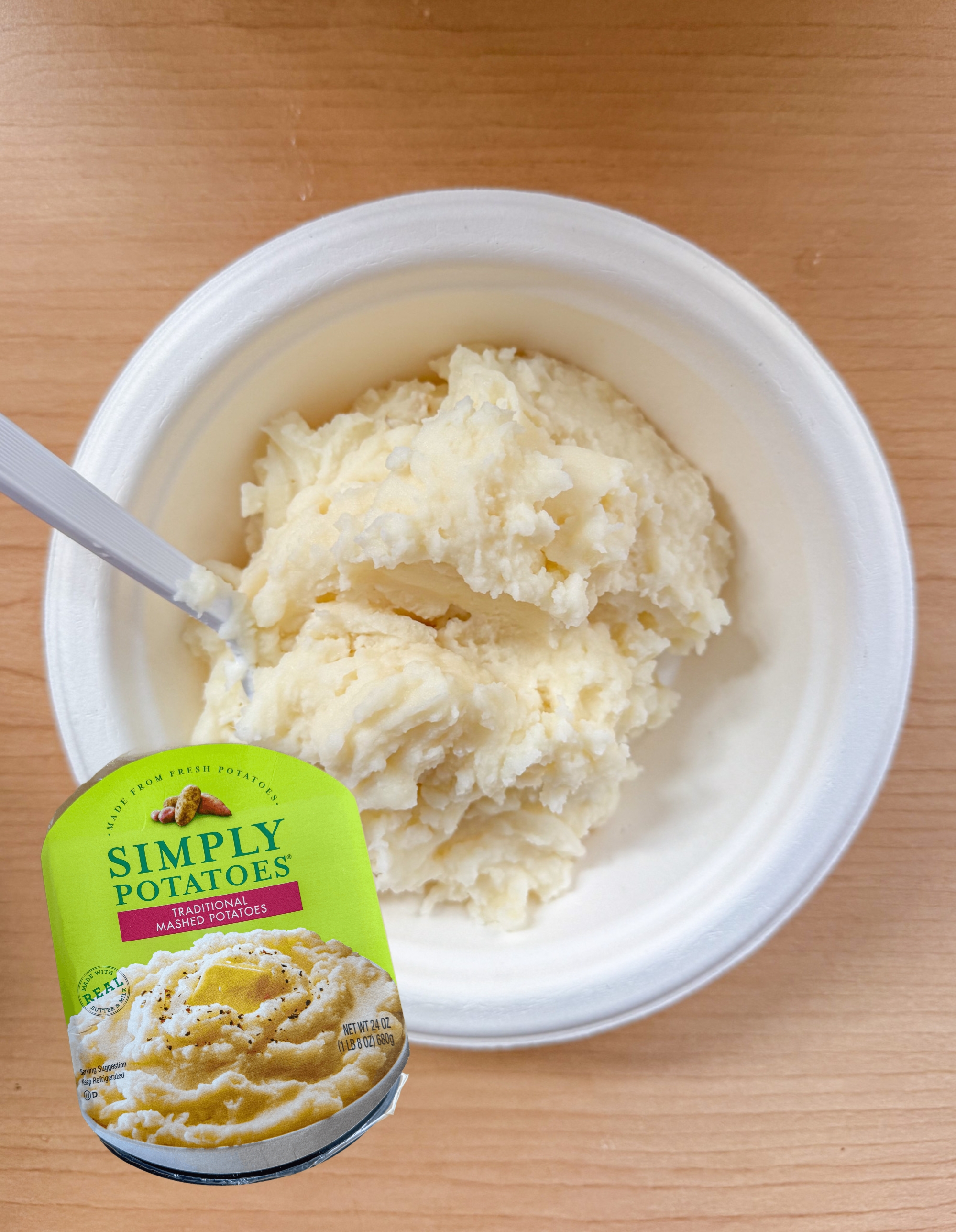 Mashed potatoes in a white bowl on a wooden table with a plastic spoon