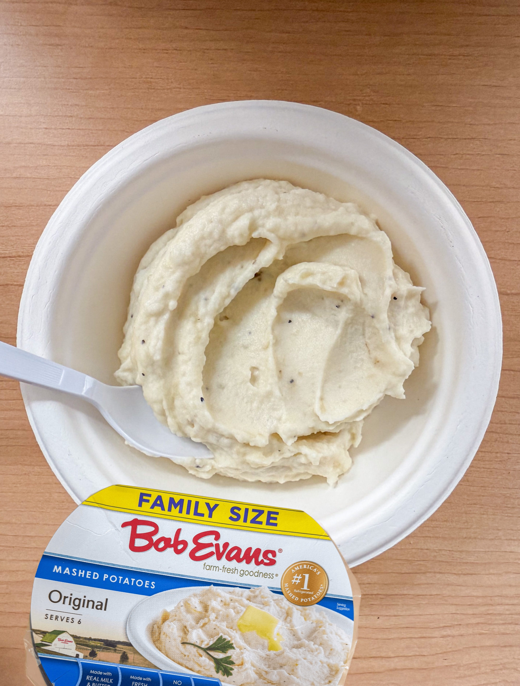 A bowl of creamy mashed potatoes with a plastic spoon on a wooden surface