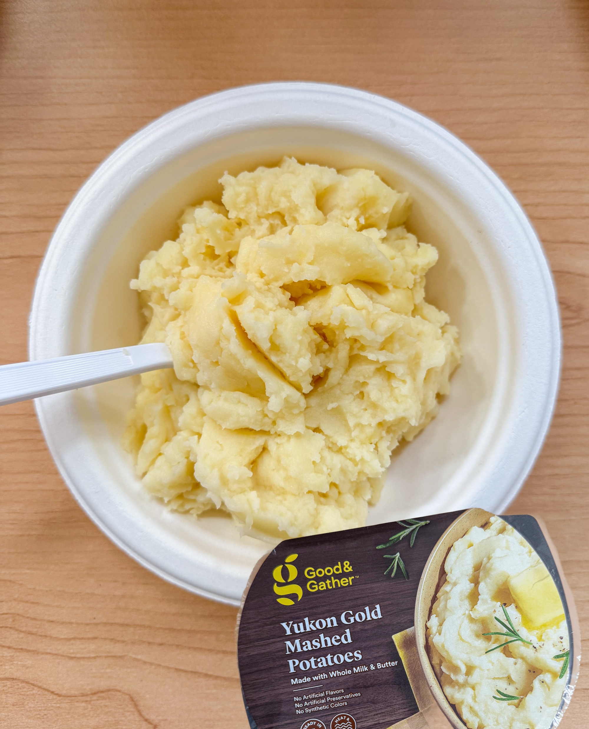 Mashed potatoes in a white bowl on a wooden surface with a plastic spoon