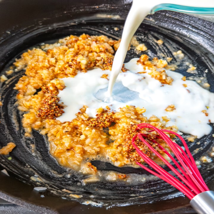Milk being poured over sautéed minced garlic in a pan with a red whisk
