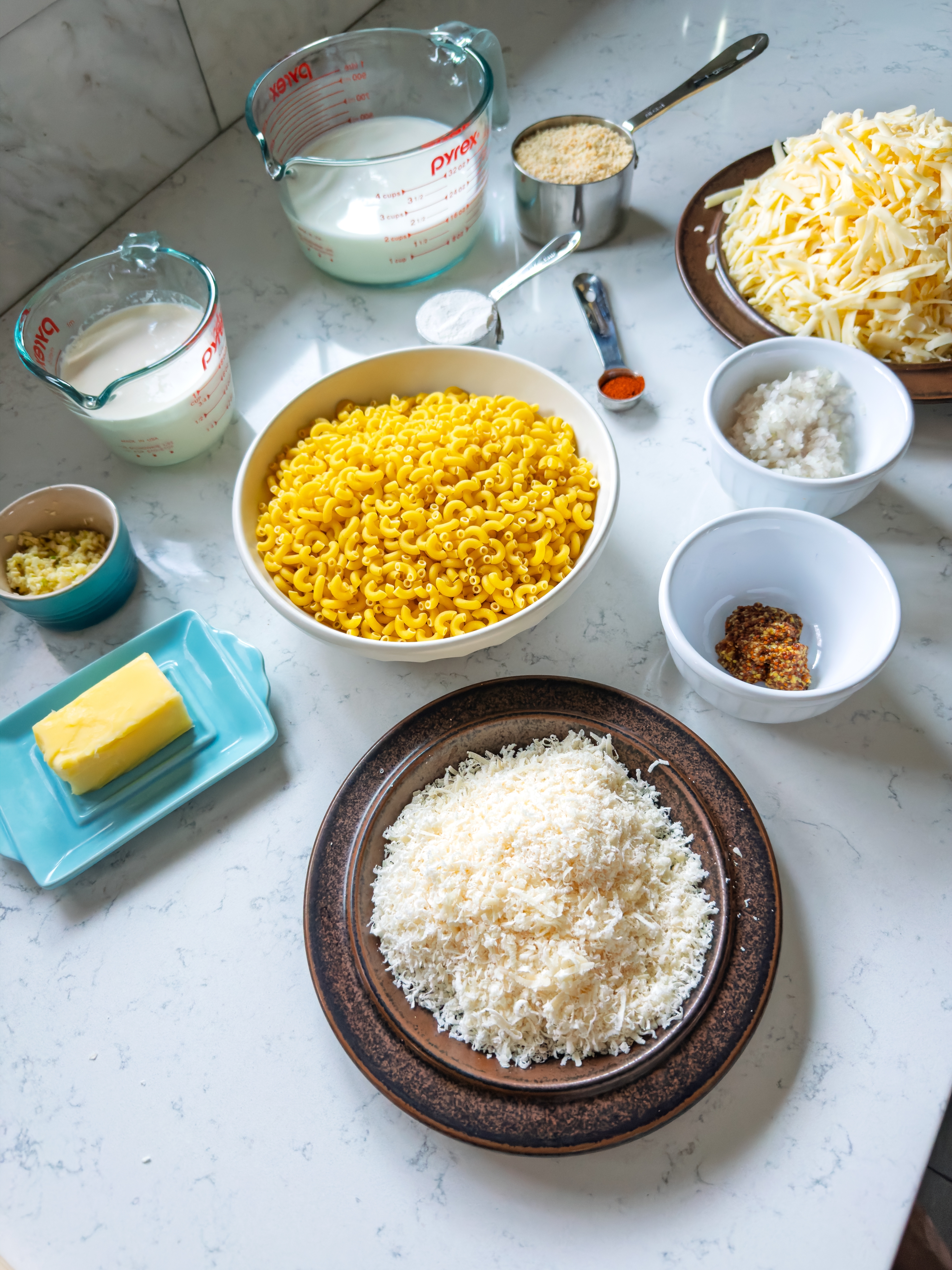 Ingredients for mac and cheese recipe including macaroni, grated cheese, butter, milk, spices, and breadcrumbs on a kitchen counter